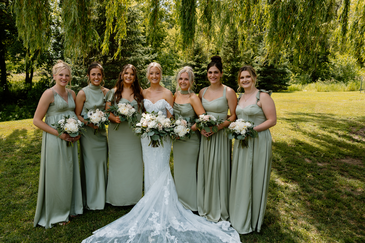 Bride poses with six bridesmaids in sage green gowns holding white bouquets under willow trees.