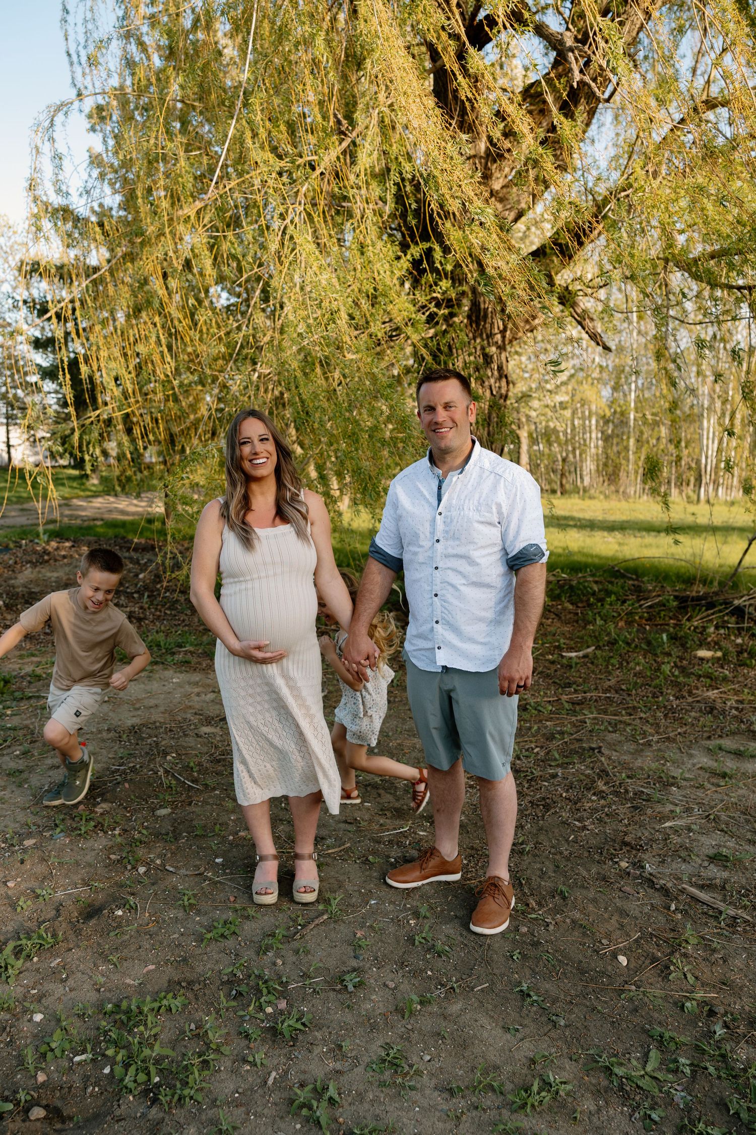 A family stands together under a willow tree on a sunny afternoon in a natural outdoor setting.