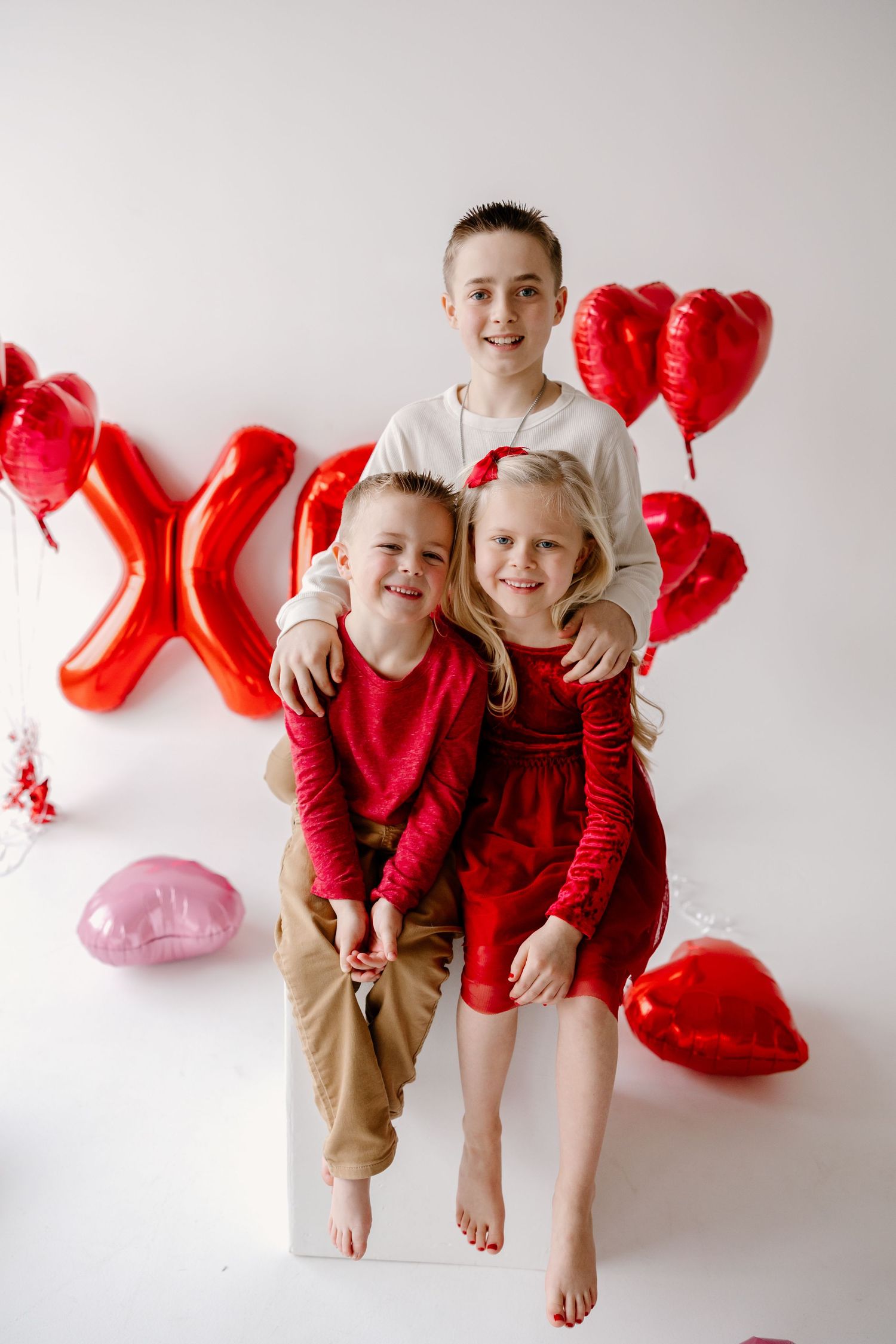 Three children pose together on white background with red and pink heart-shaped Valentine's Day balloons.