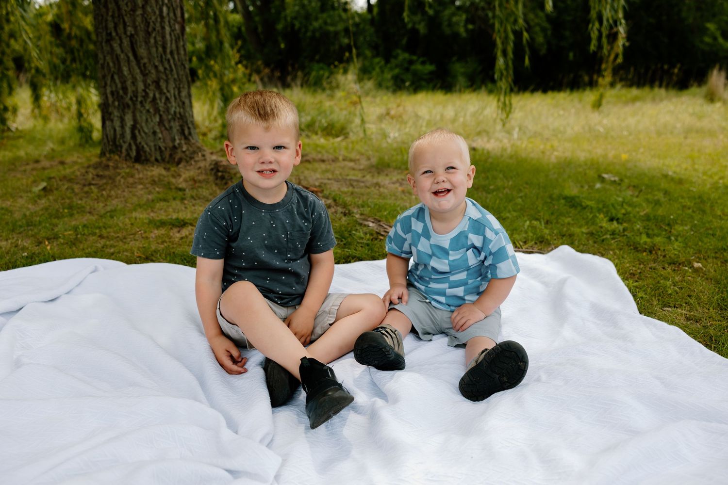 Two young children sit together on a white blanket outdoors on a sunny summer day in a grassy area.