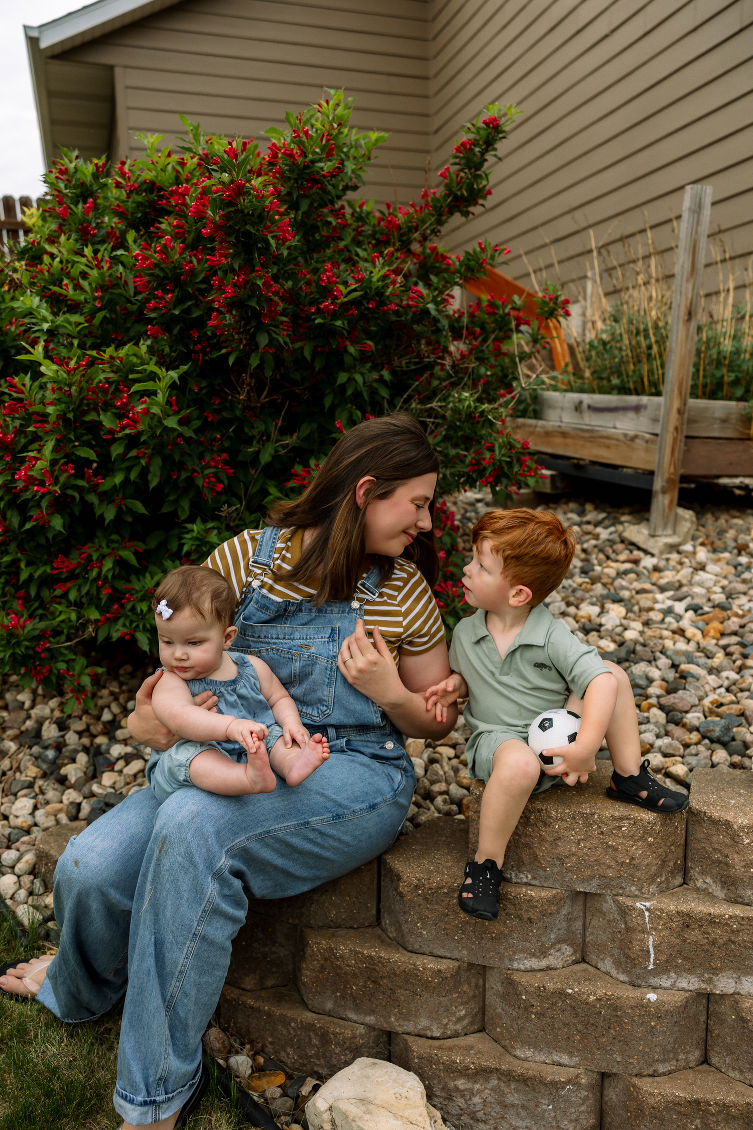 A mother in denim overalls sits on stone steps with her baby girl and toddler son in a garden.