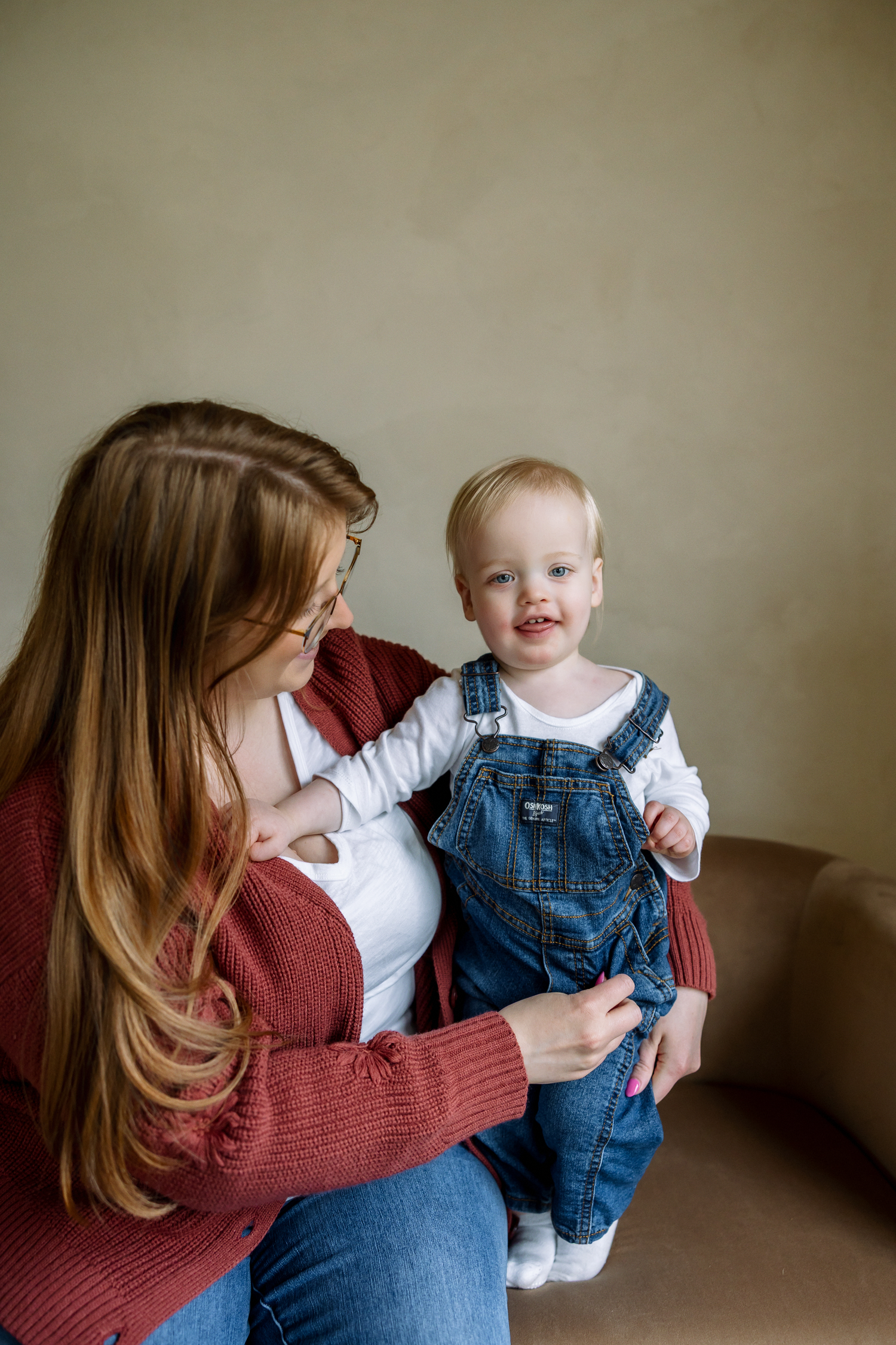 A smiling toddler in denim overalls sits on a woman's lap in a warm indoor setting.