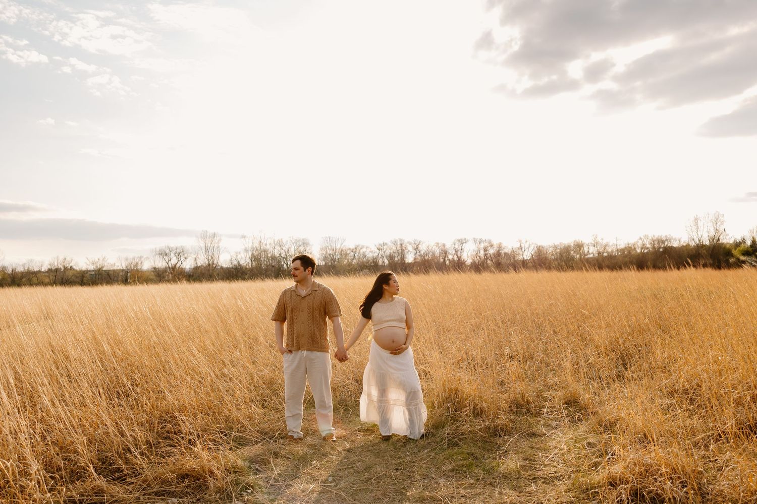 A couple holds hands while walking through a golden wheat field at sunset on a dreamy autumn afternoon.