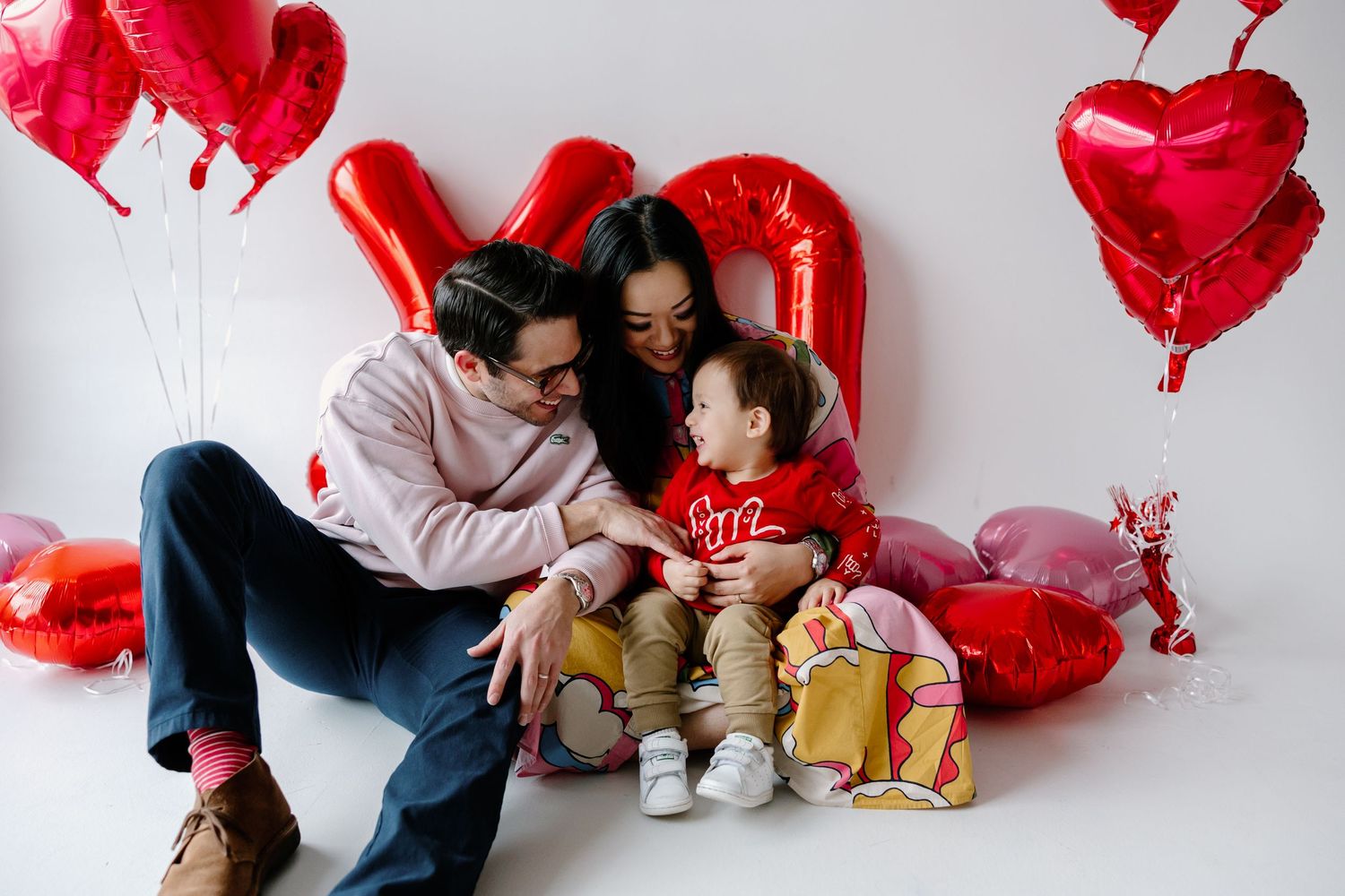 A family sits together on the floor surrounded by red heart-shaped balloons for Valentine's Day photos.