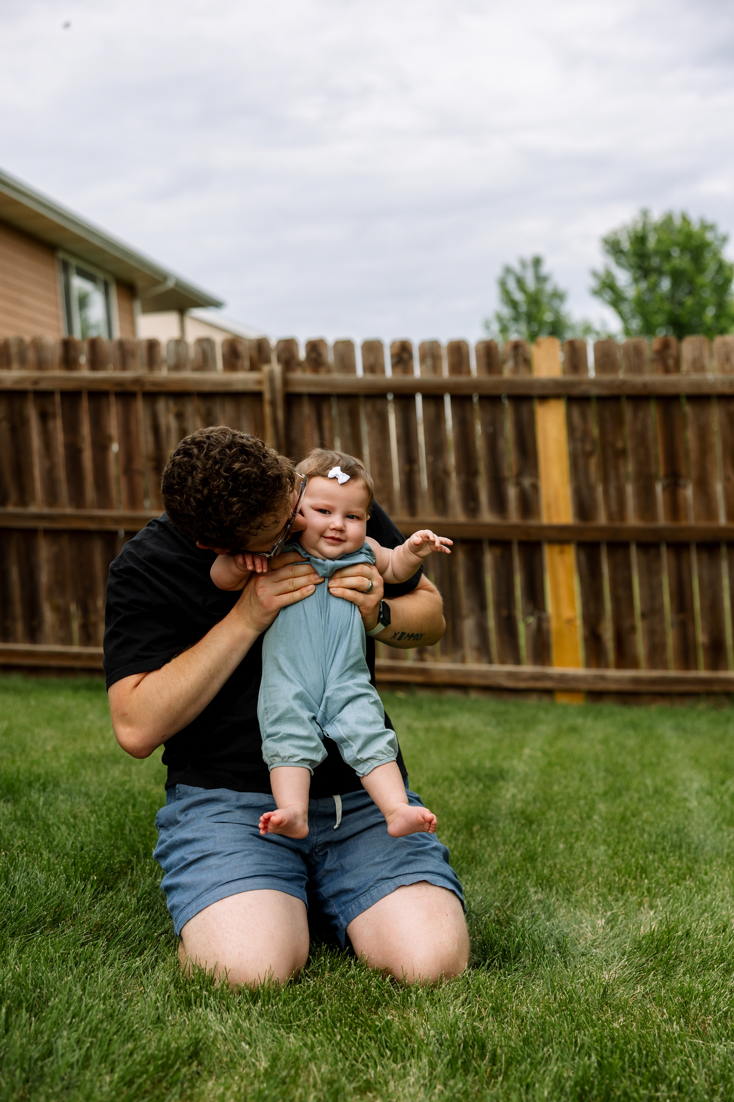 Parent kneeling on green grass holding and kissing a smiling baby in a light blue outfit outdoors.