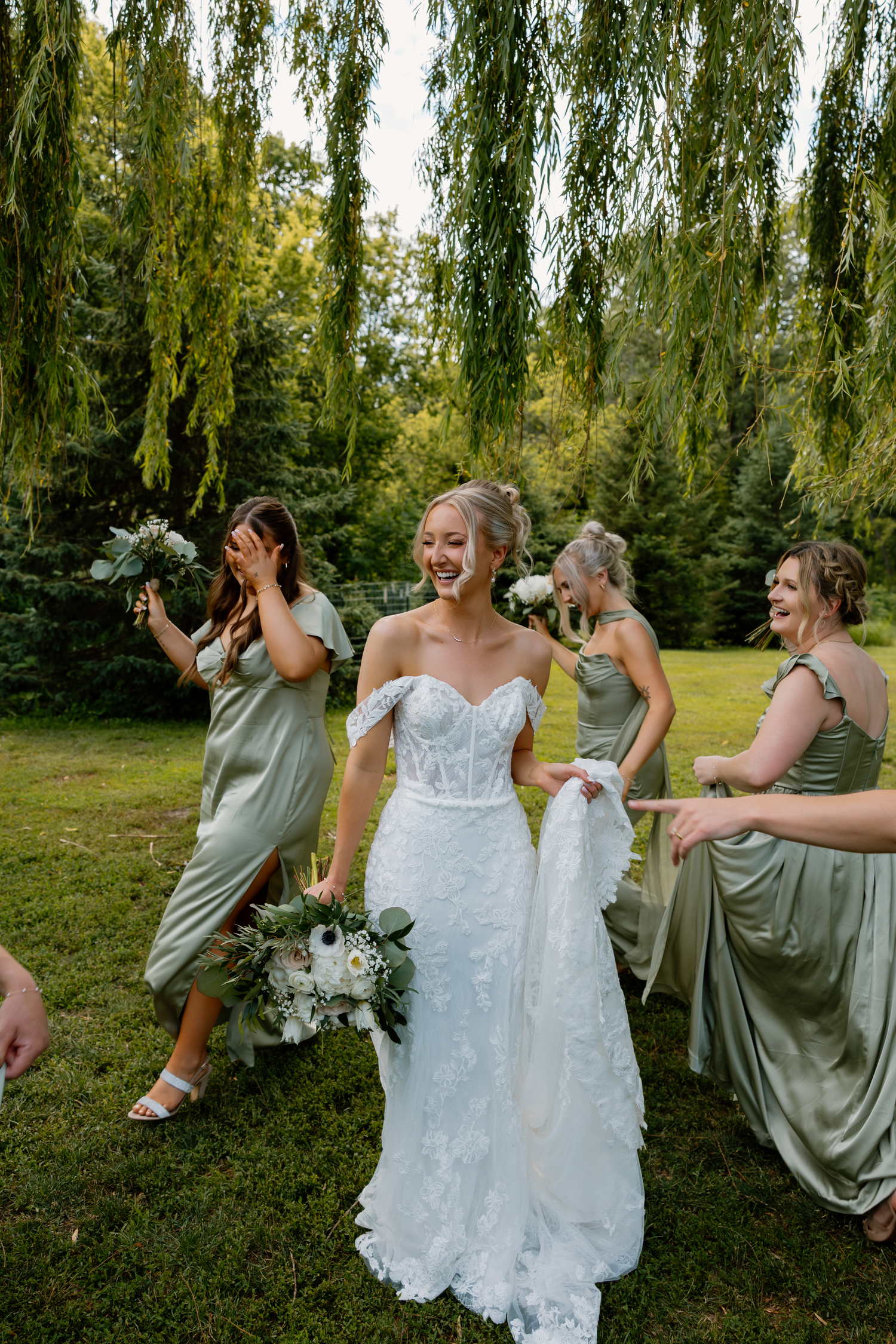 Bride in white lace gown laughs with bridesmaids in sage green dresses under weeping willow trees.