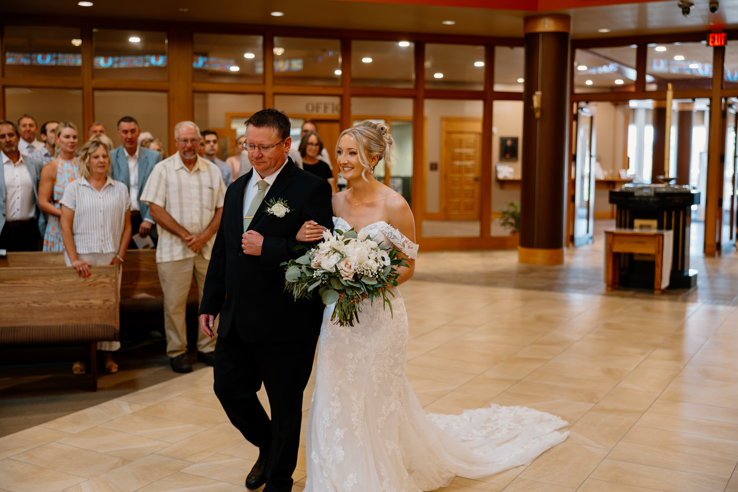 Bride in lace gown walks down aisle with escort holding white floral bouquet at church wedding.