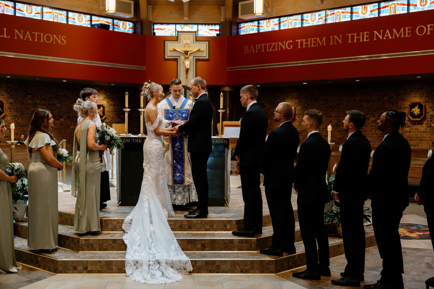 Bride and groom exchange vows at church altar surrounded by wedding party during elegant ceremony.