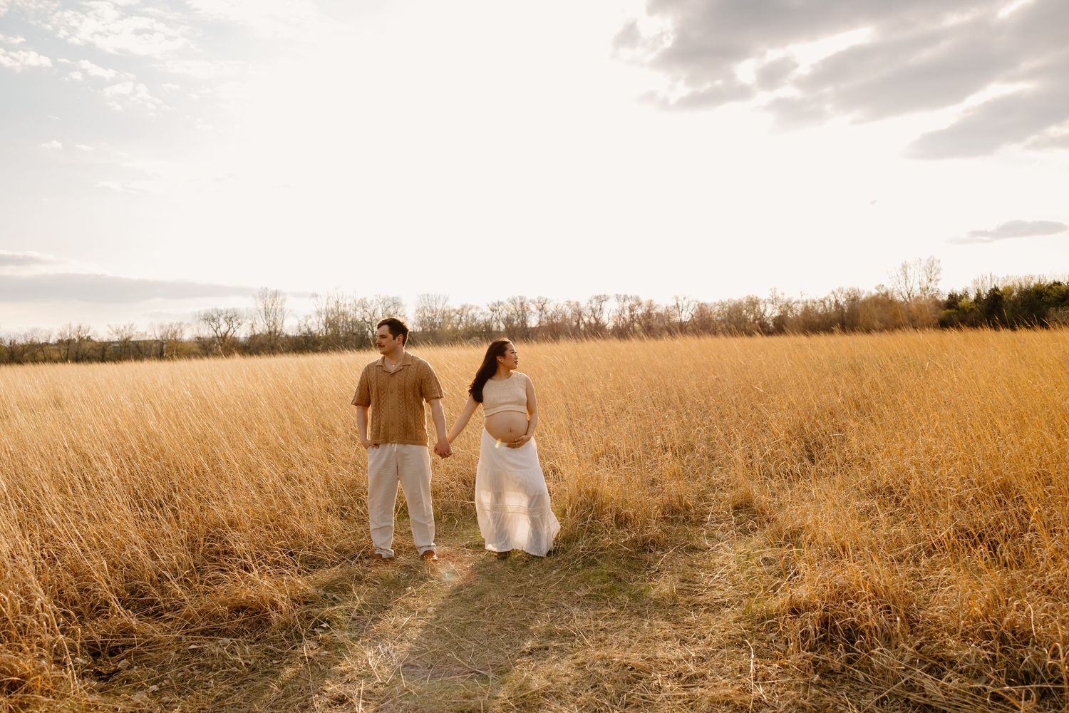 Two people walk hand in hand along a dirt path through golden wheat fields at sunset.