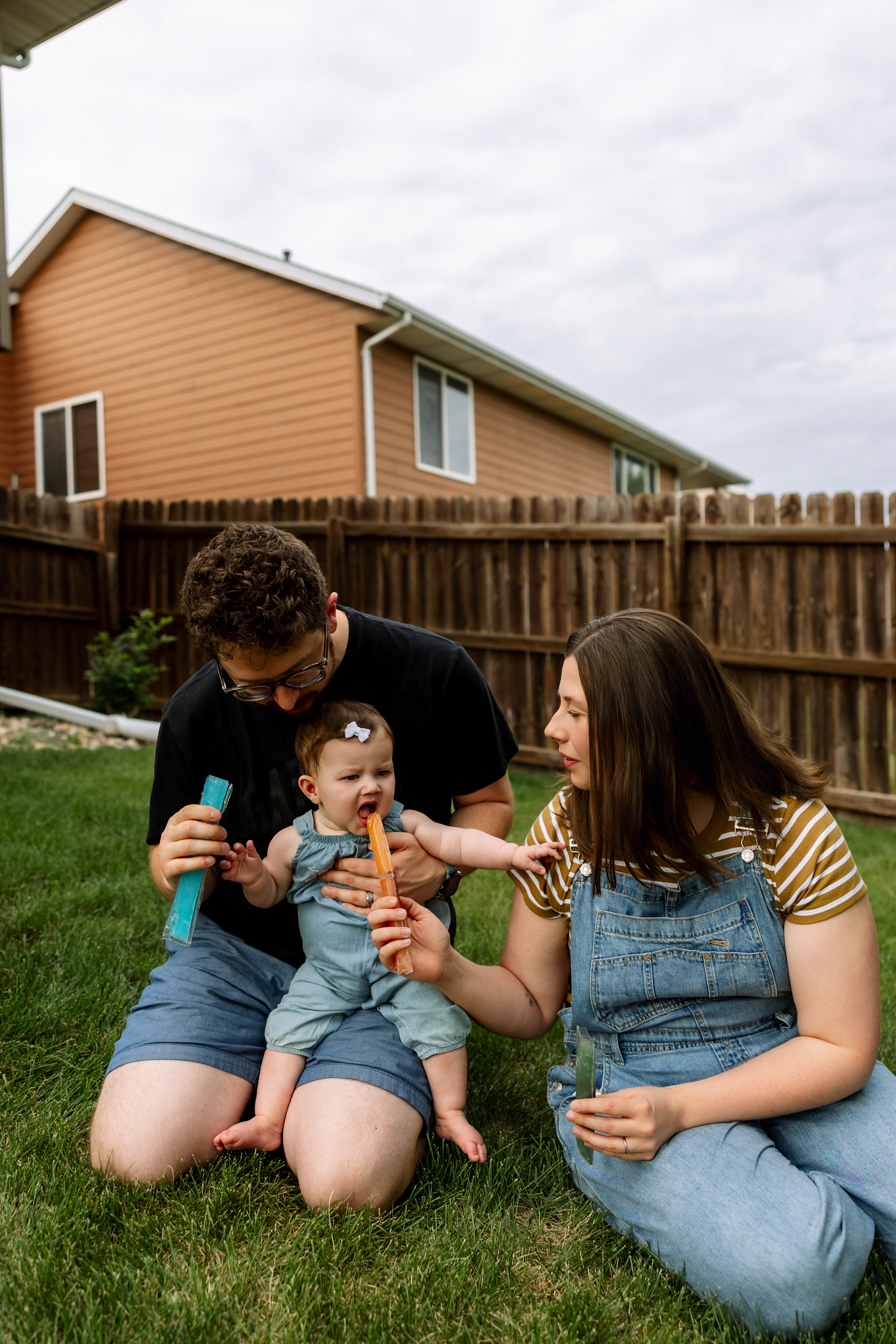 Parents sitting on grass feeding baby popsicles in fenced backyard.