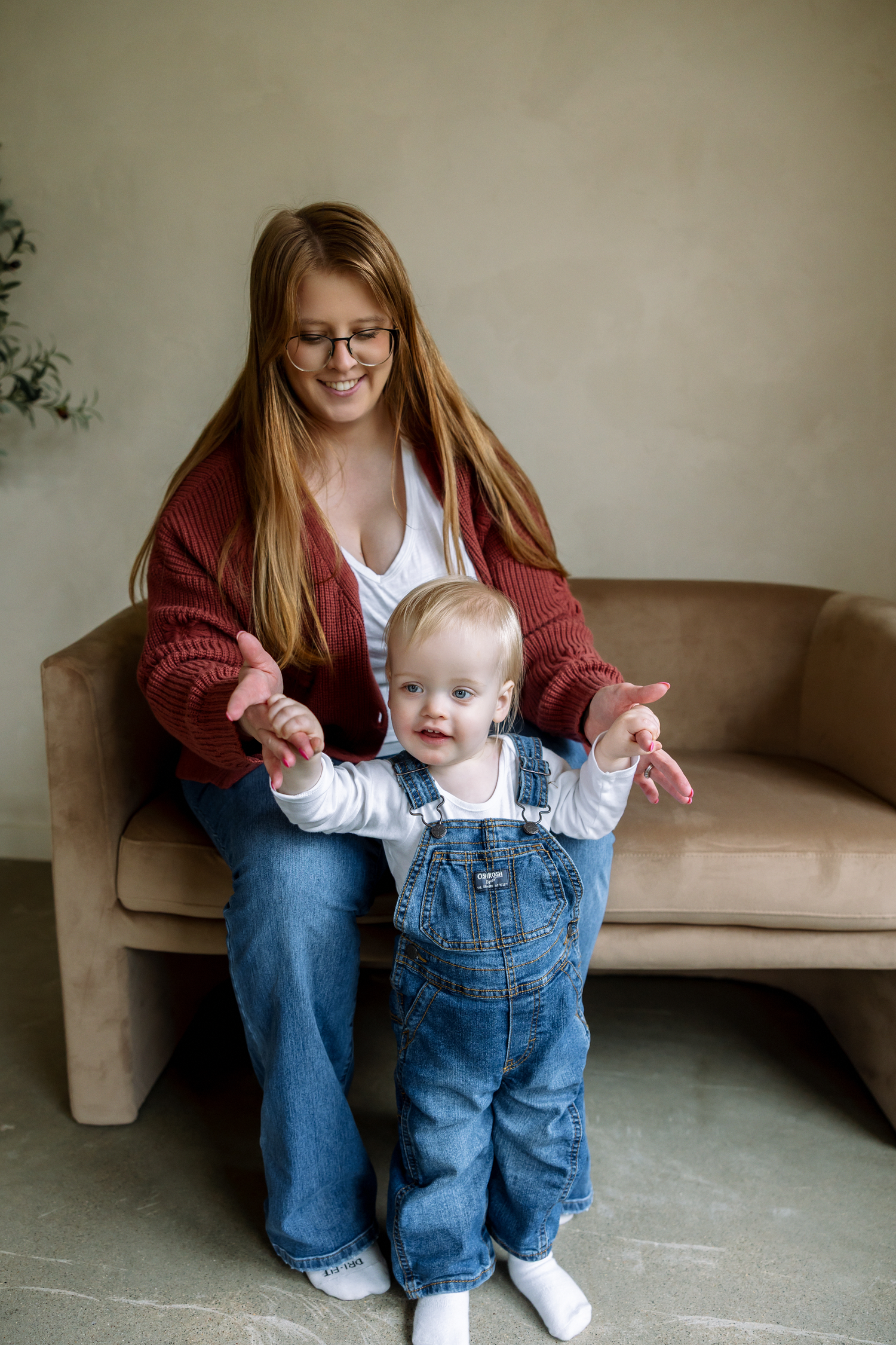 A smiling redhead mother in glasses helps her baby in denim overalls stand on her lap on a beige couch.