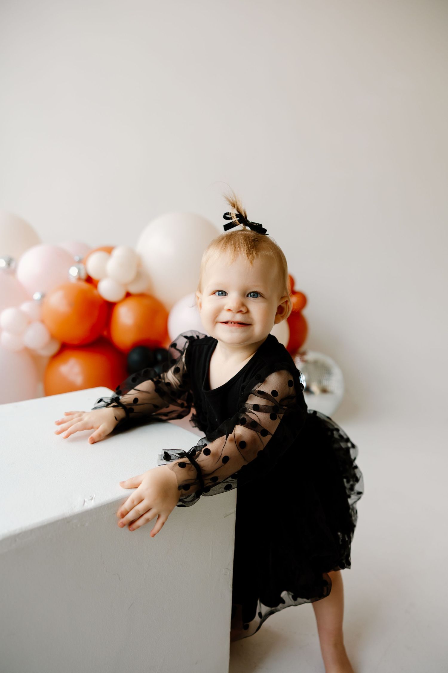 A small child in a black dress poses playfully near white and orange balloons against a light colored wall.