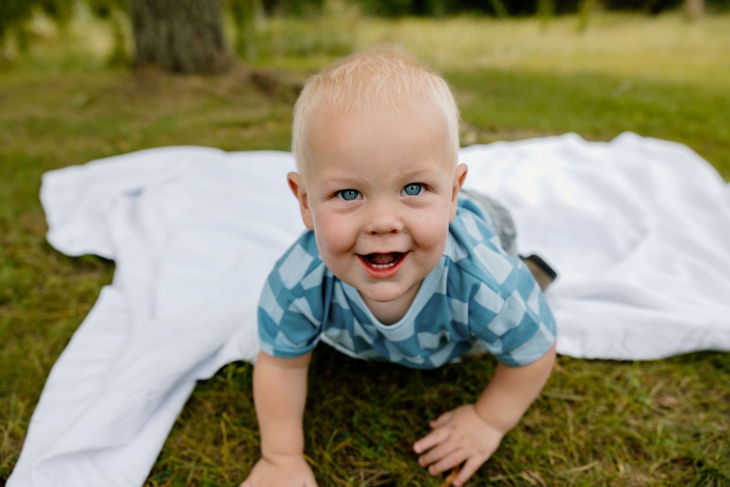 Happy child in blue plaid shirt crawling on white blanket outdoors with natural background.