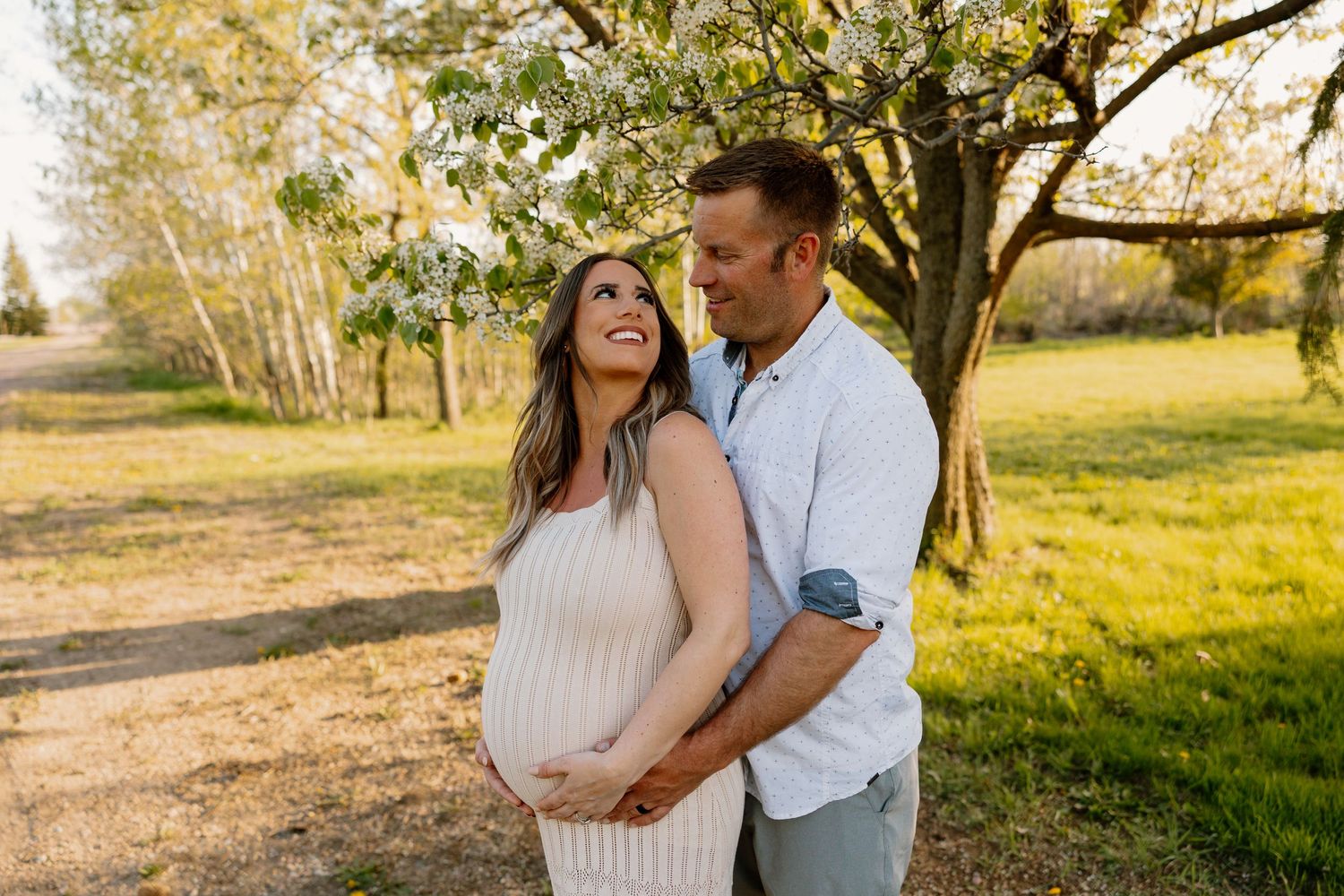 A couple embraces in a sunlit park during a maternity photoshoot with fall trees and golden light in the background.