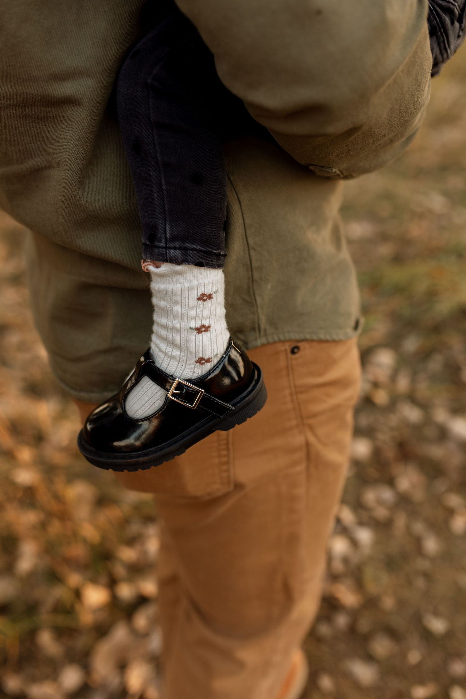Child wearing black Mary Jane shoes and white ribbed socks being carried by an adult in tan corduroy pants outdoors.
