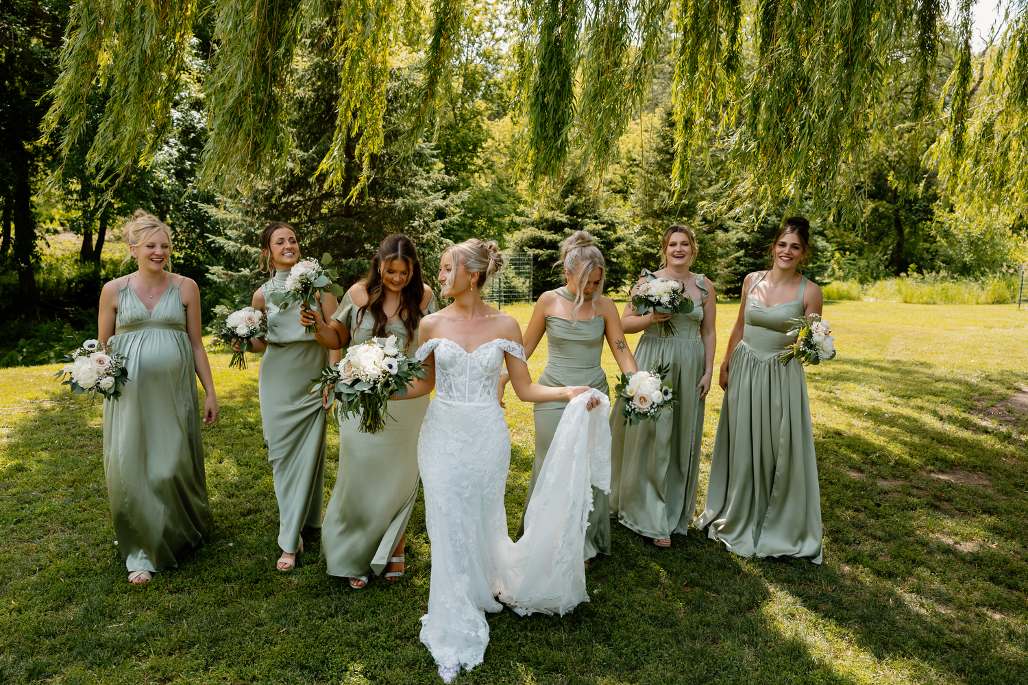 Bride in white gown with bridesmaids in sage green dresses walking under weeping willow trees.