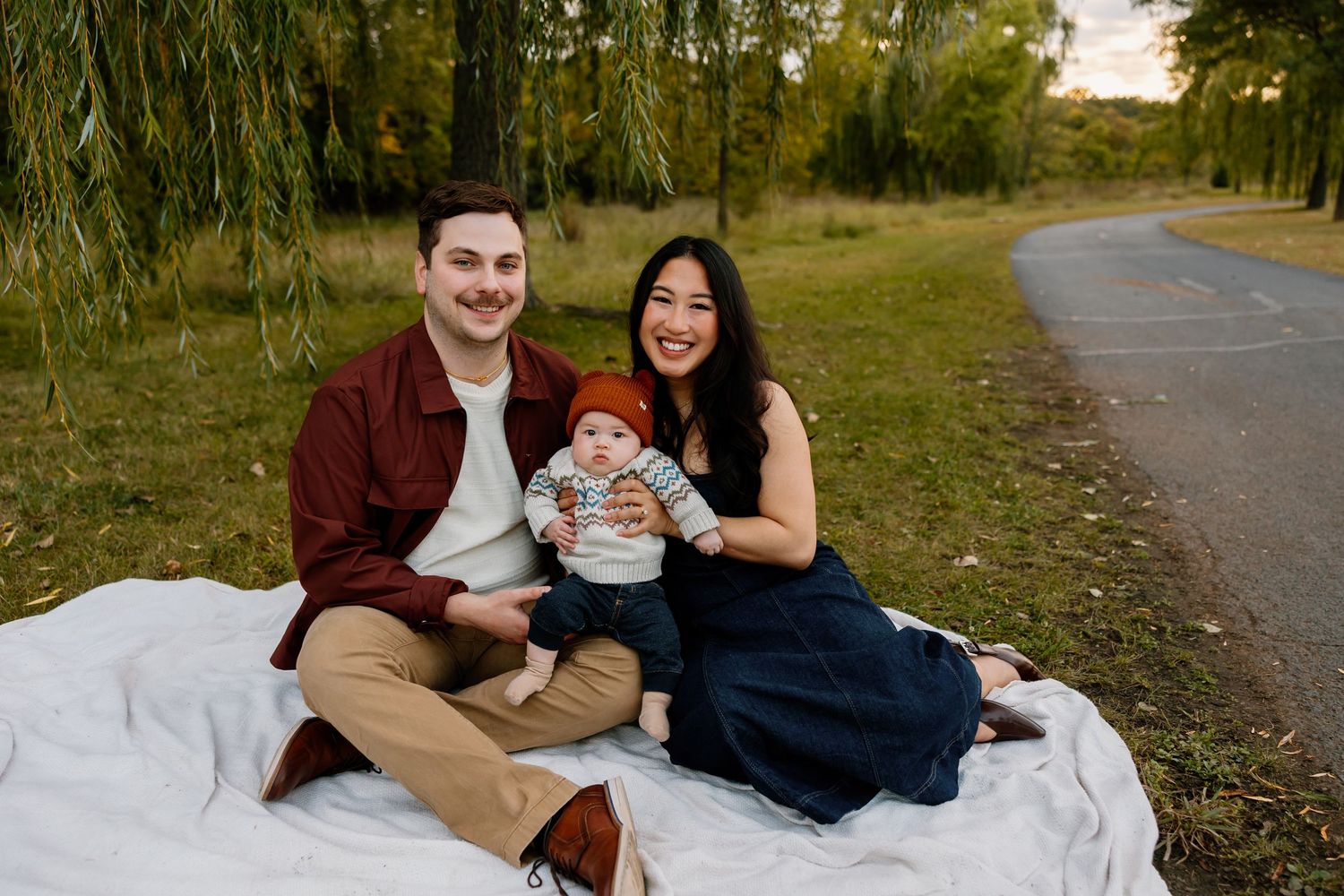 Family of three sitting on blanket outdoors, parents smiling with baby in fall park setting.