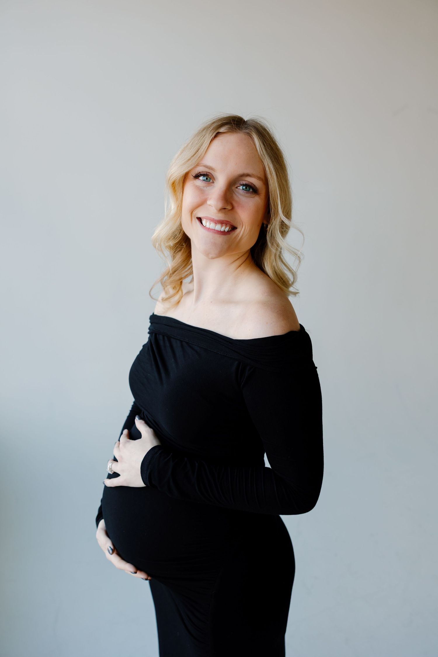 A pregnant woman in a black sweater poses against a white wall during a maternity photoshoot. Riley Ro Photo Sioux Falls, SD