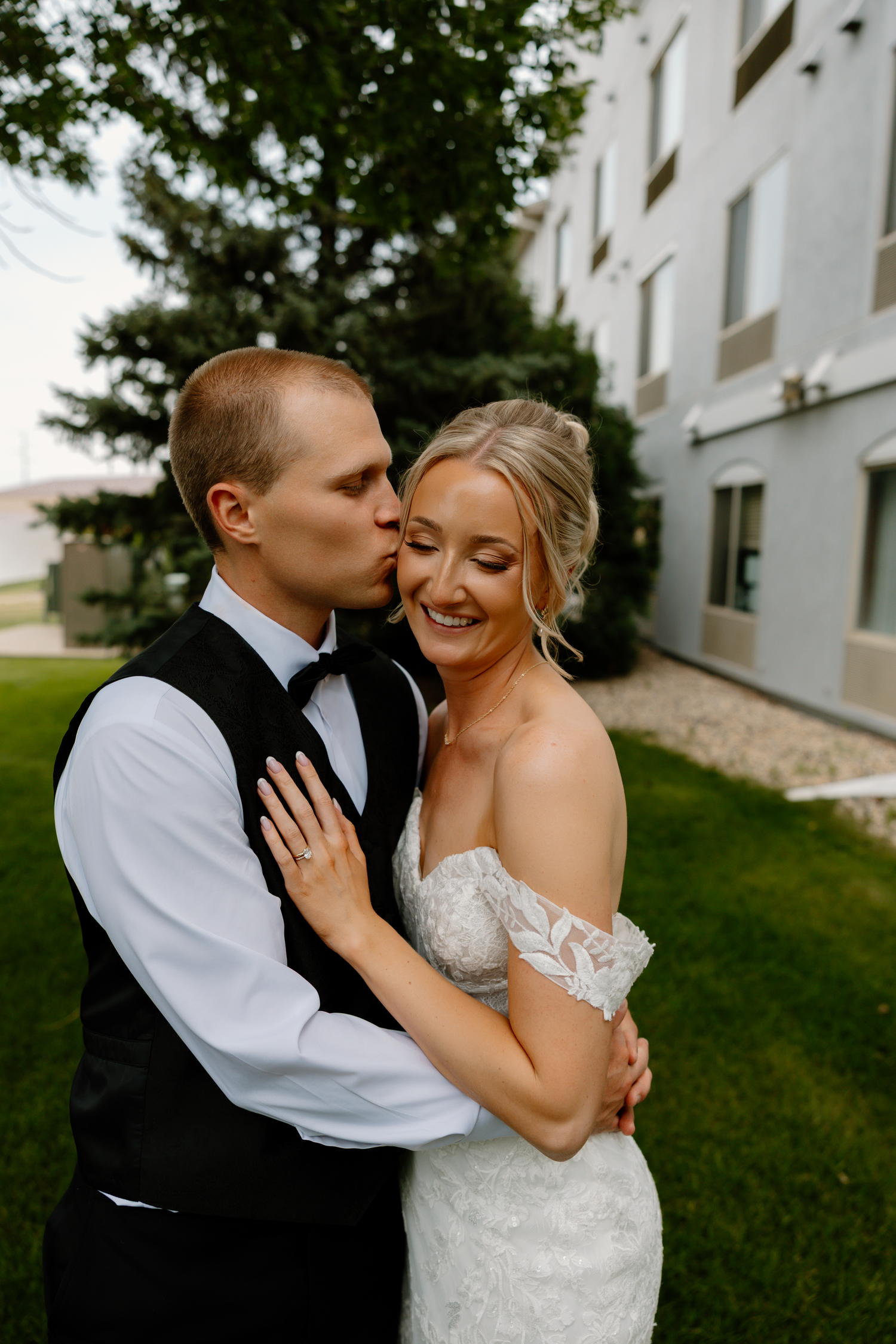 Groom in black vest kisses bride on cheek as she smiles in lace off-shoulder wedding dress outdoors.