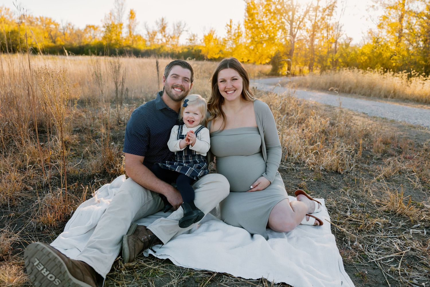 Happy family maternity photo outdoors, parents sitting on blanket in golden field with toddler, fall foliage backdrop.