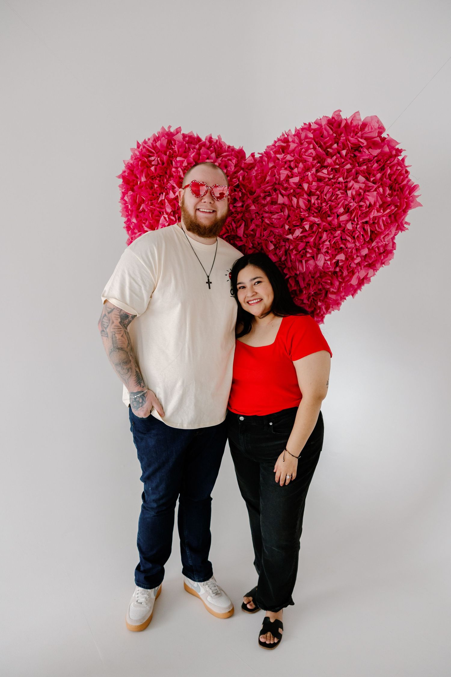 A couple poses together in front of a large pink floral heart decoration against a white studio backdrop.