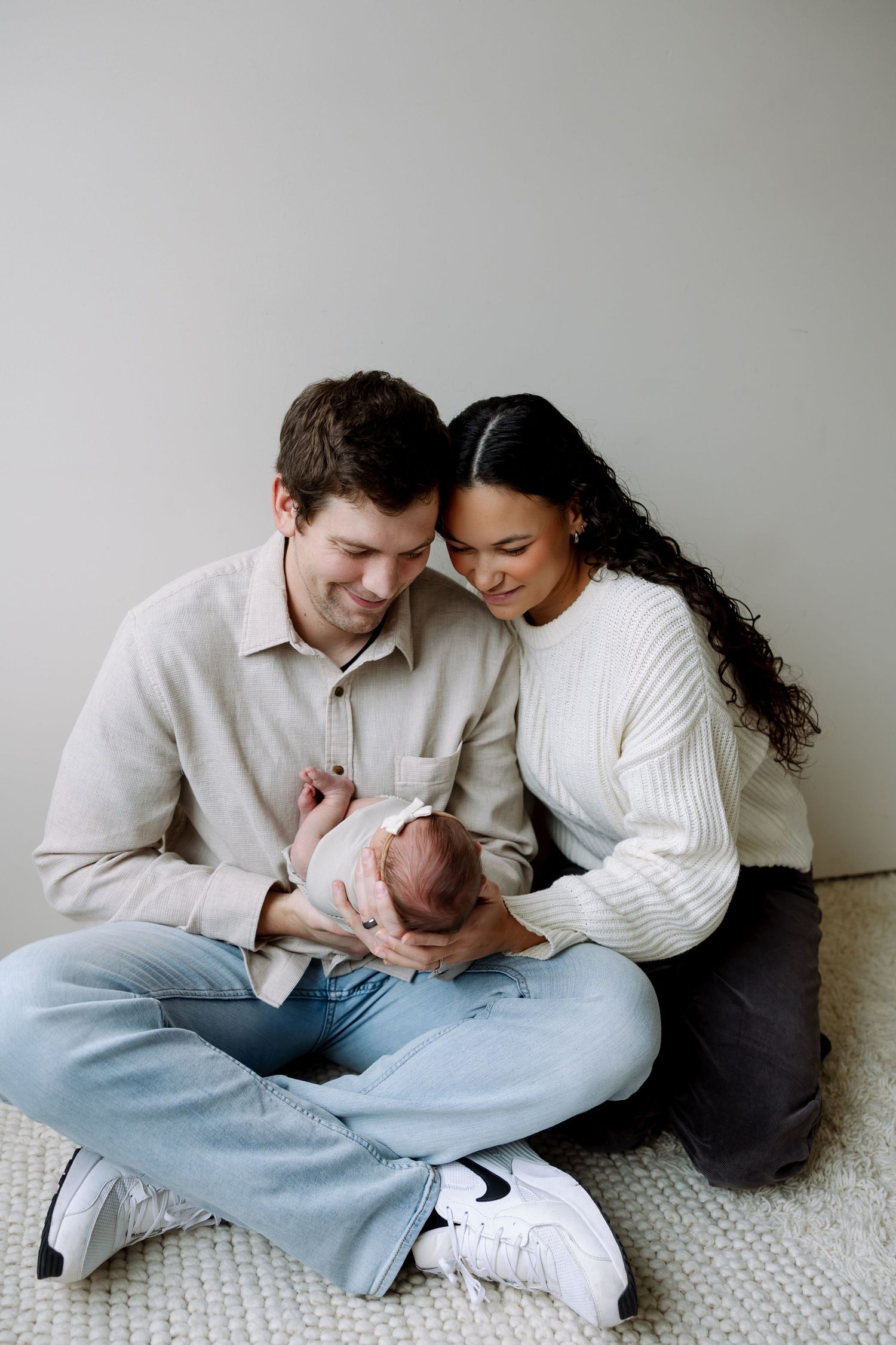 A couple in white sweaters and jeans sits intimately together on a light carpet in a minimalist setting.
