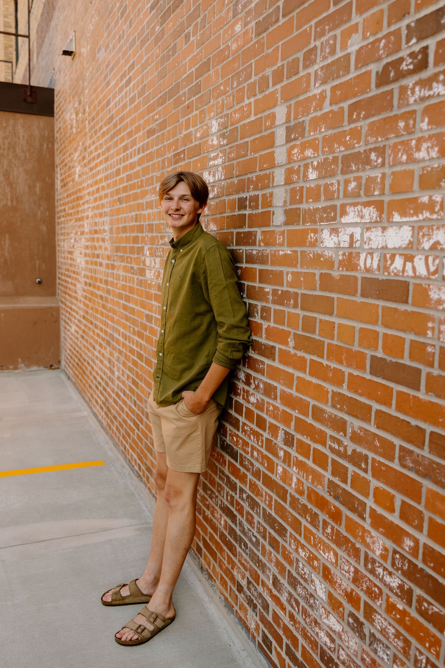 A casual outfit featuring khaki shorts and an olive shirt is modeled against a rustic brick background.