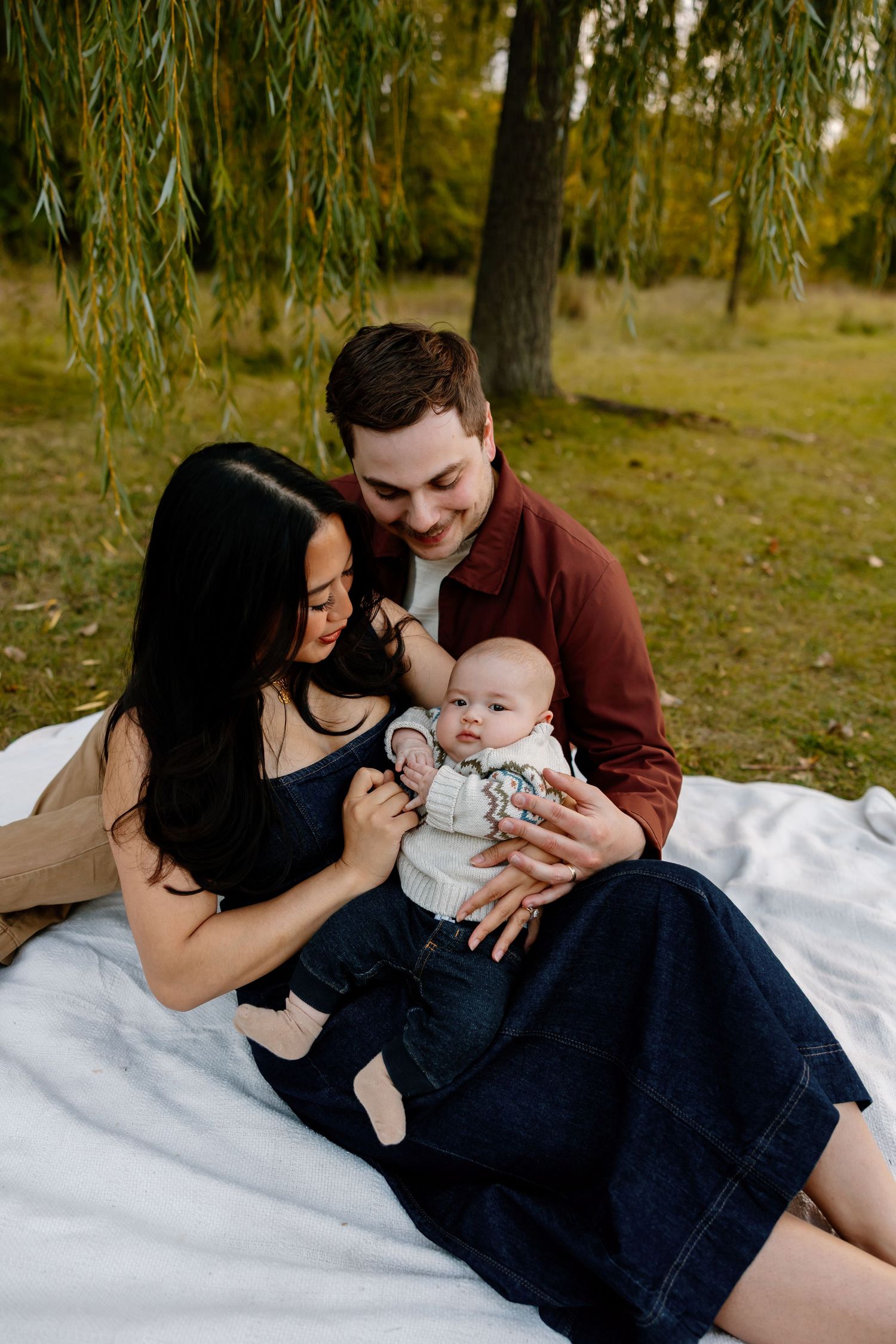 A family sits on a white blanket under willow trees in a park during autumn, sharing a tender moment with their baby. Riley Ro Photo Sioux F