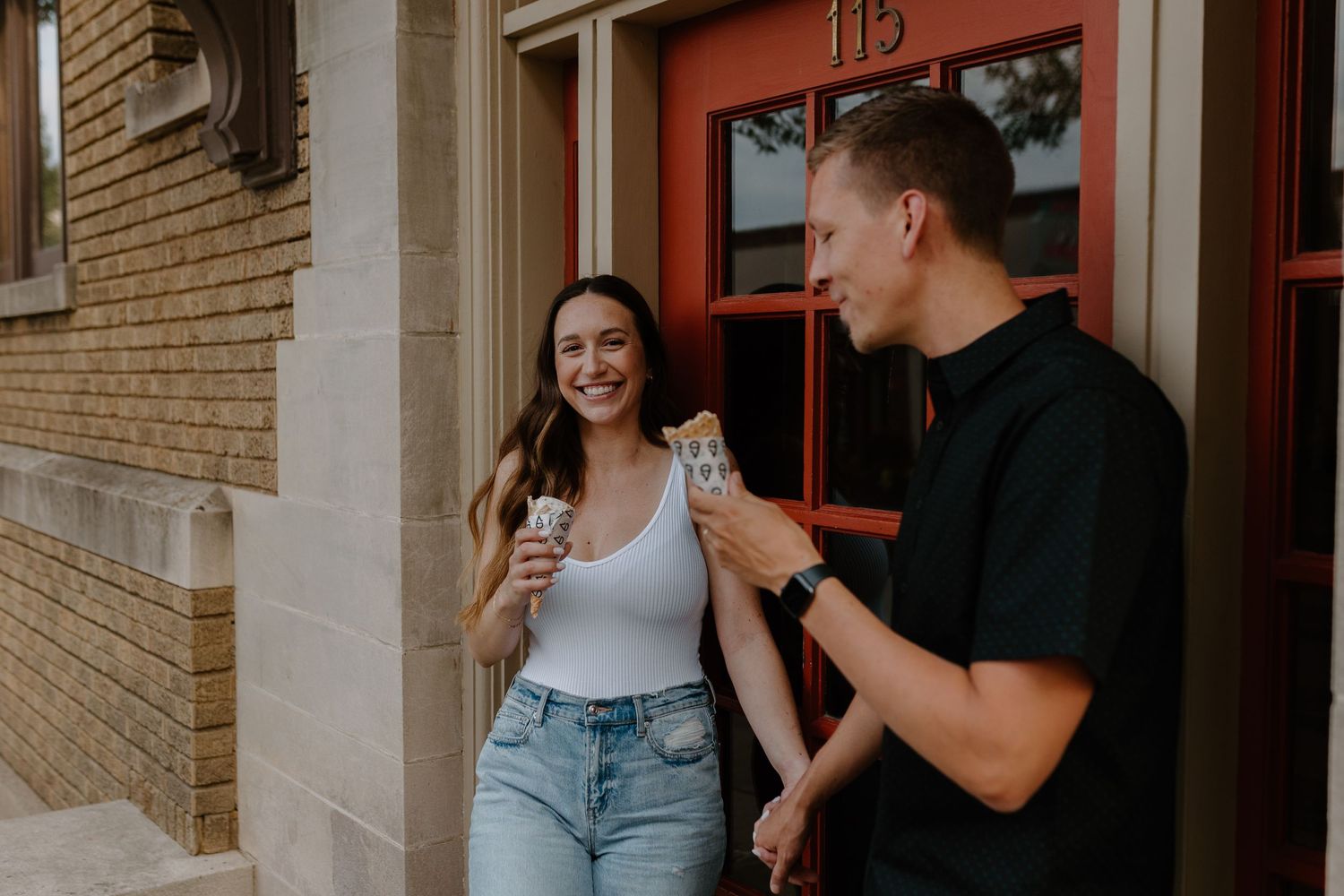 A couple enjoying drinks and laughing together outside a building with a red door.