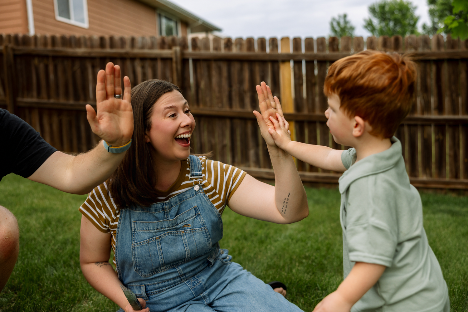 A woman in denim overalls laughs while high-fiving a red-haired boy in a sunny backyard.