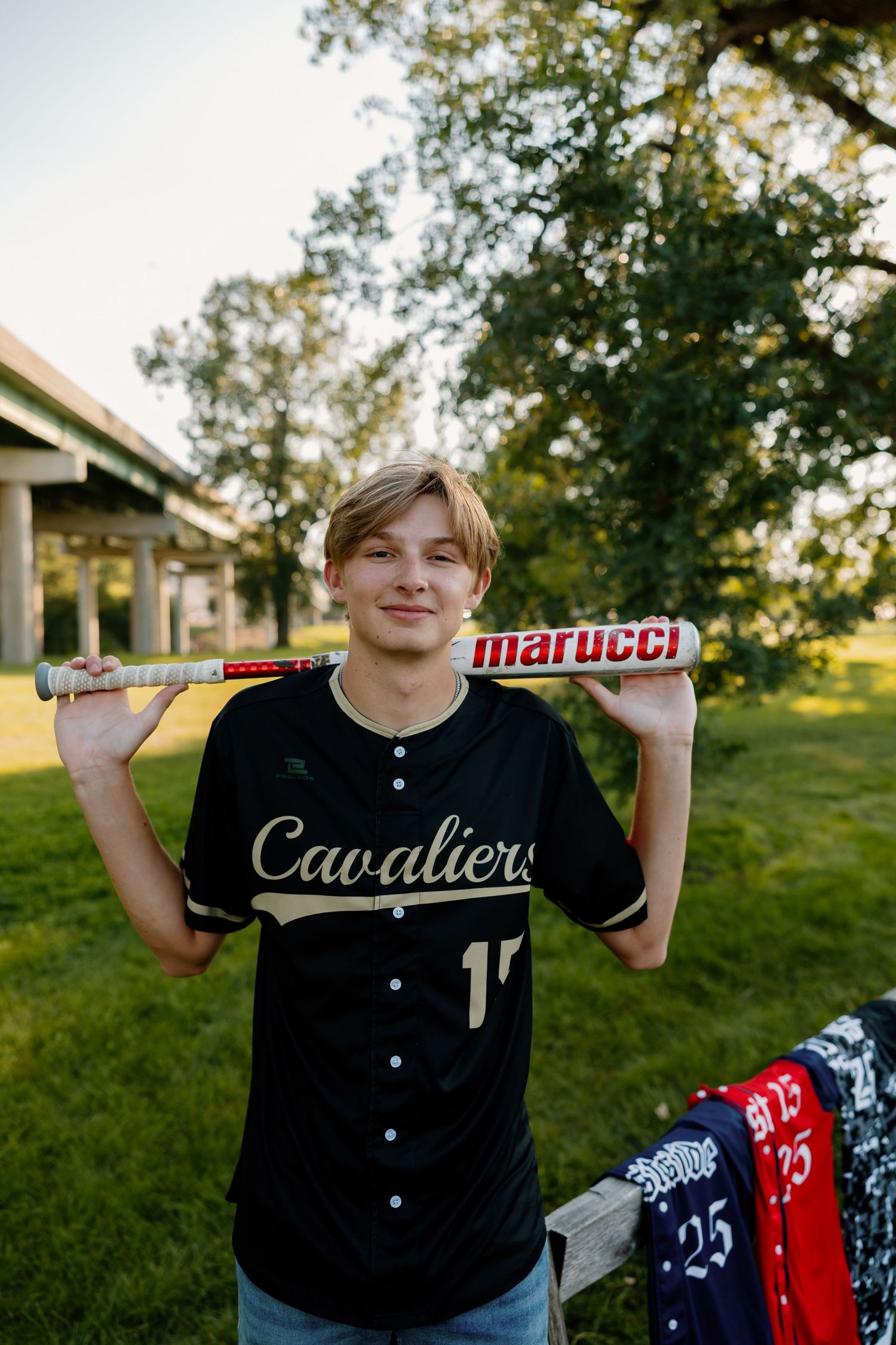 Baseball player in black uniform poses with Marucci bat and jerseys displayed.