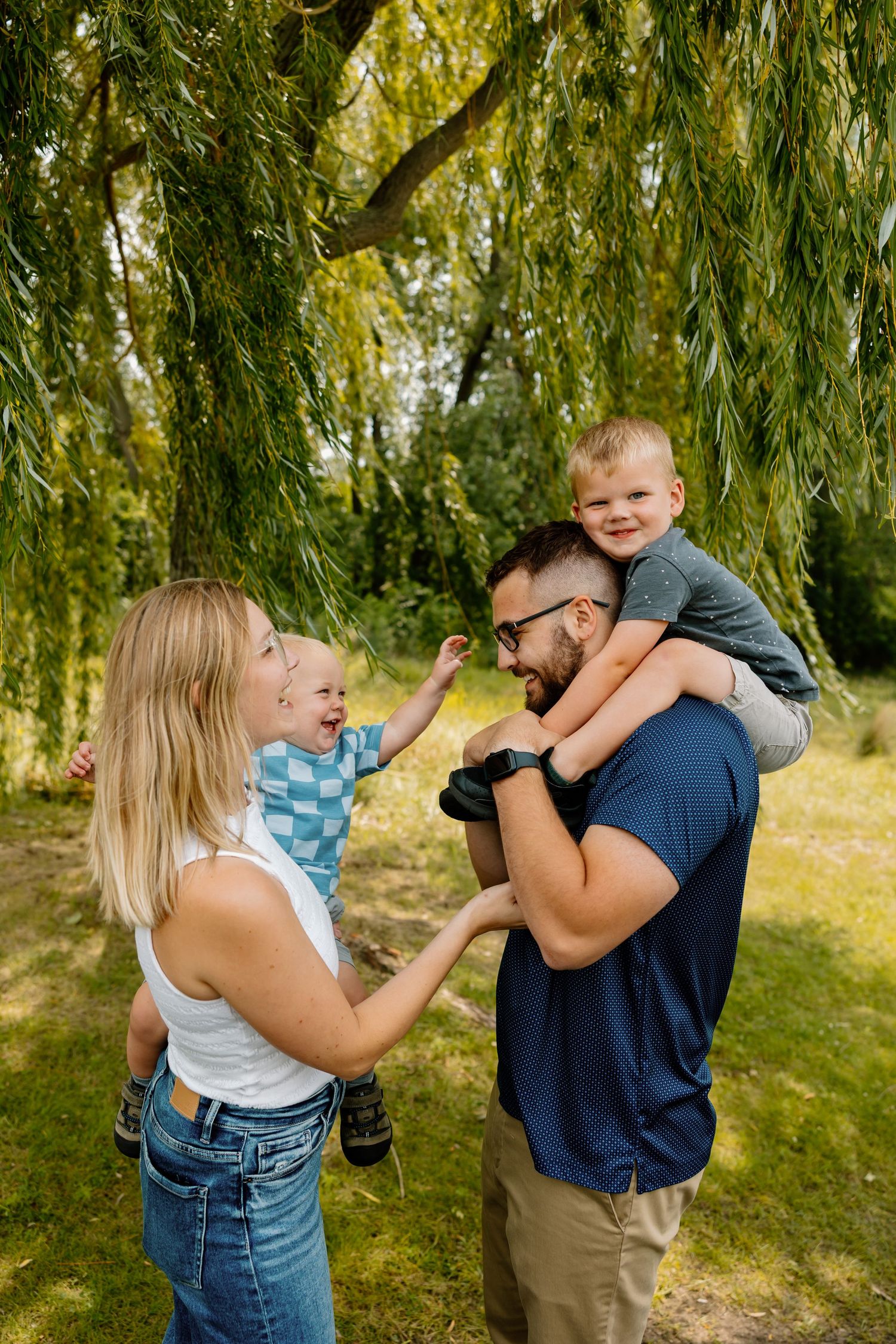 Happy family playing and laughing together under drooping willow branches in a park setting.