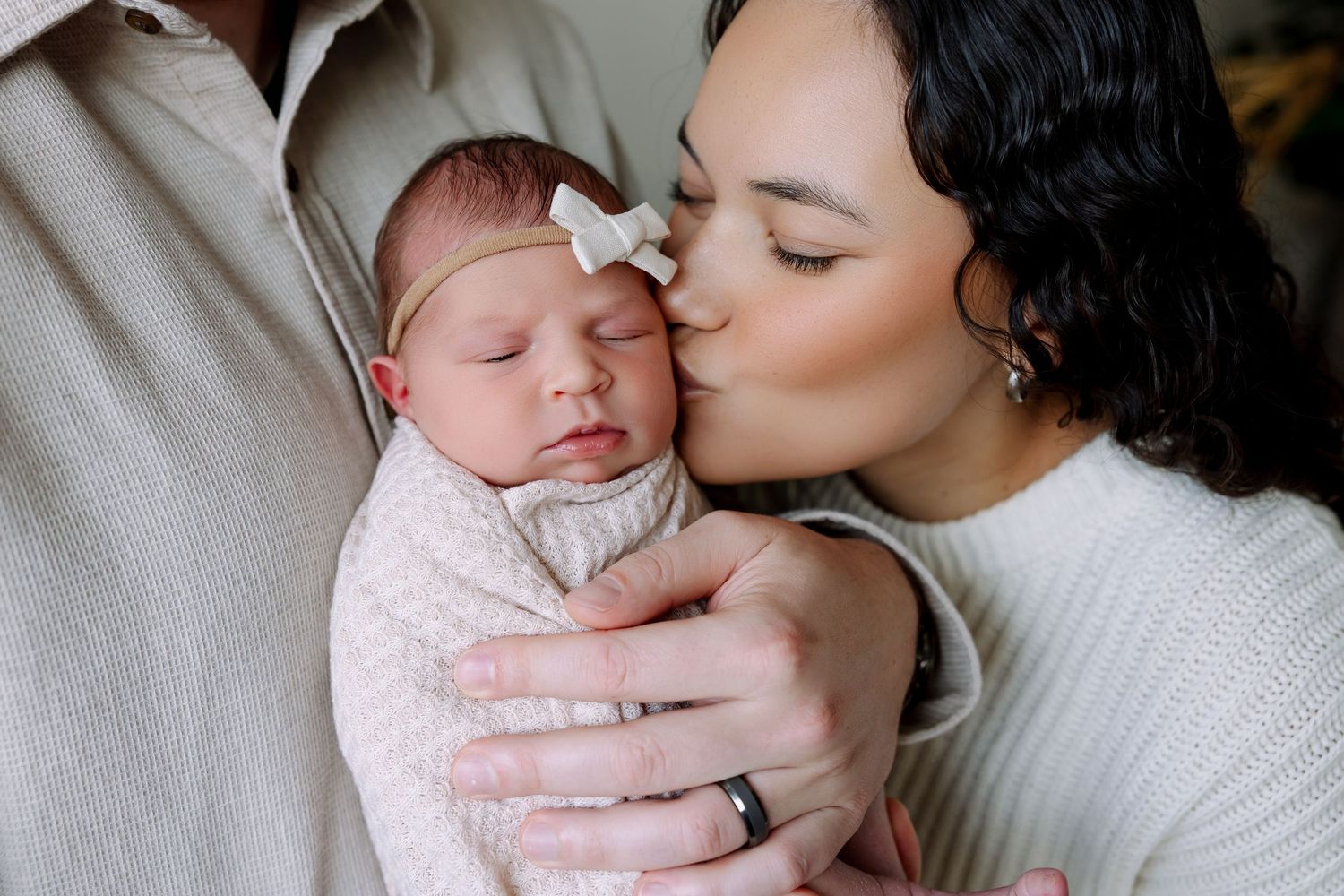 A mother tenderly kisses her sleeping newborn baby wrapped in a soft pink swaddle blanket.