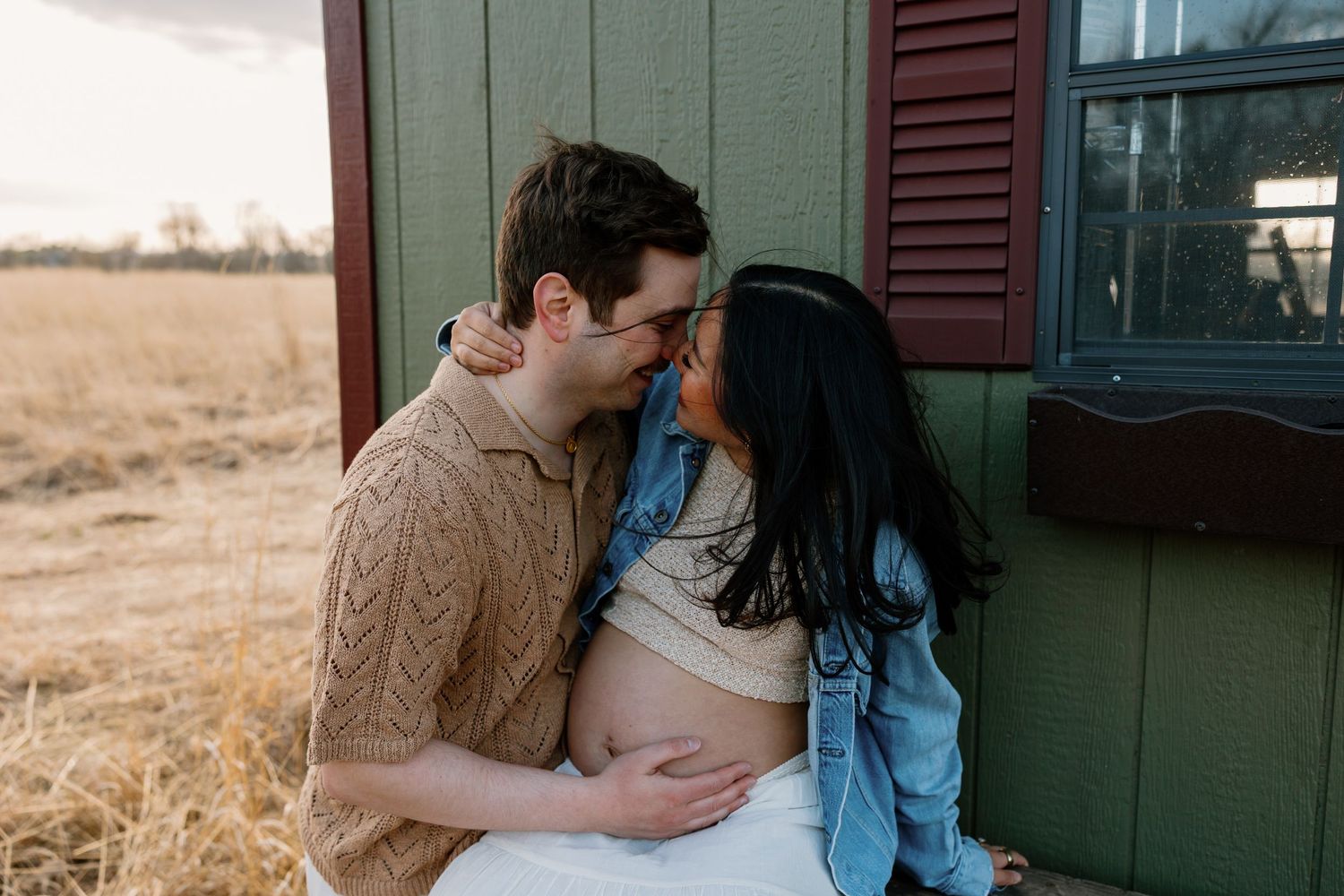A romantic couple embraces in front of a green building during sunset in a golden wheat field.