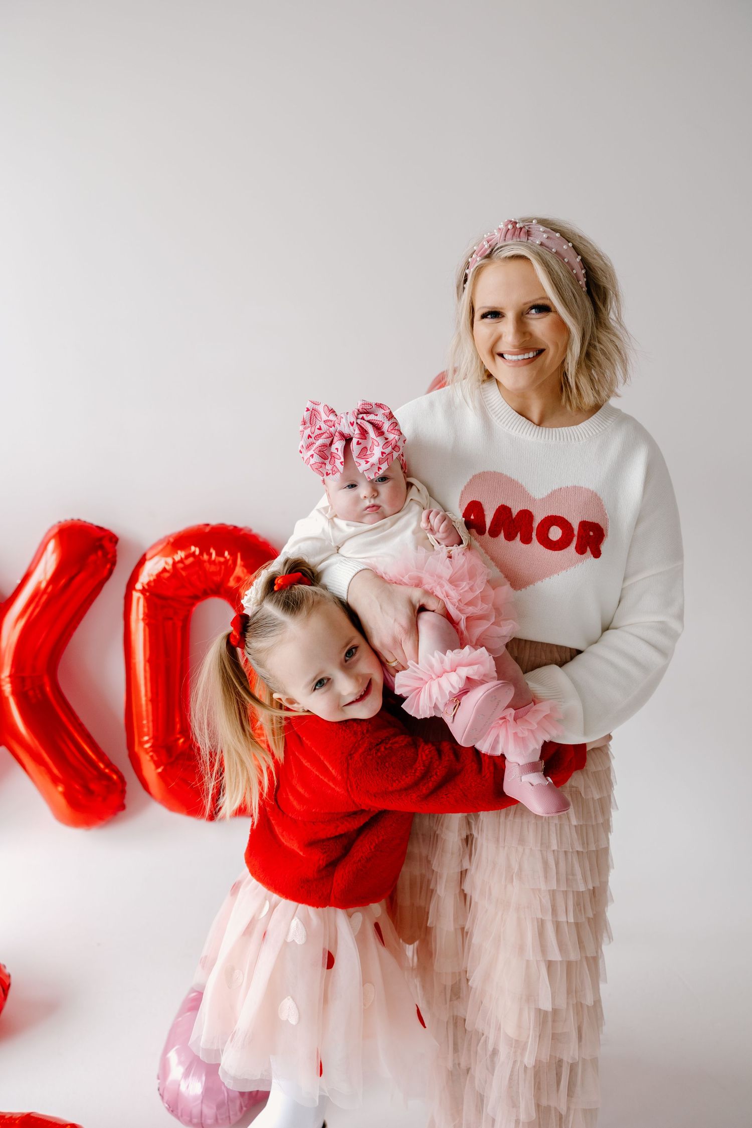 Holiday studio photo with woman in Amor sweater and tulle skirt hugging child against white wall with red balloons.