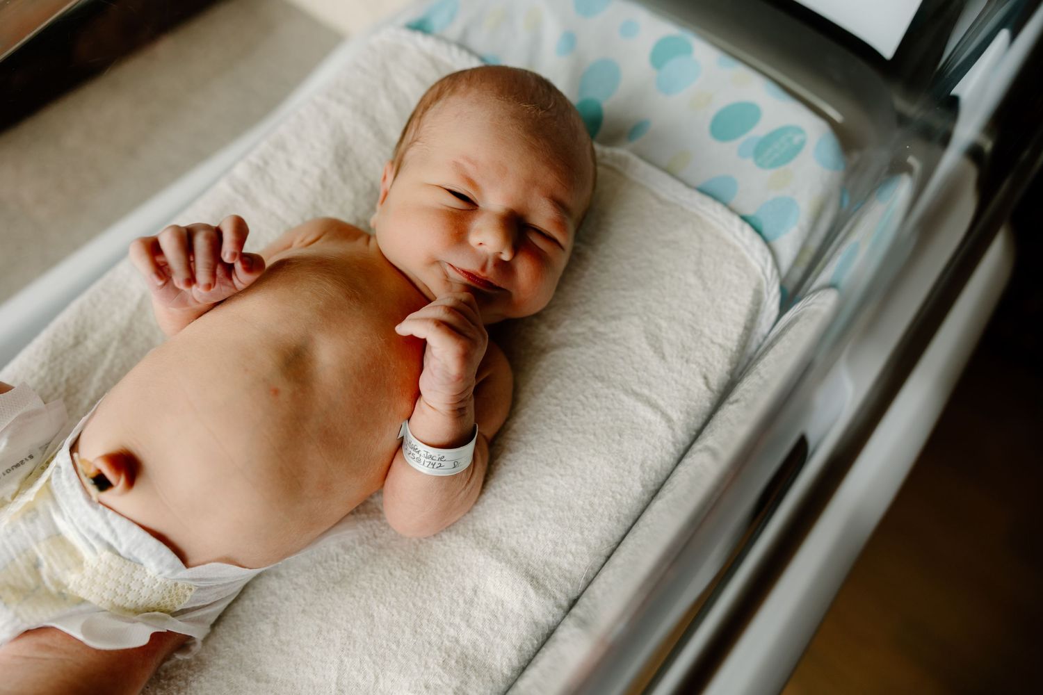 Newborn lies on white bedding in a hospital bassinet while stretching their arms.