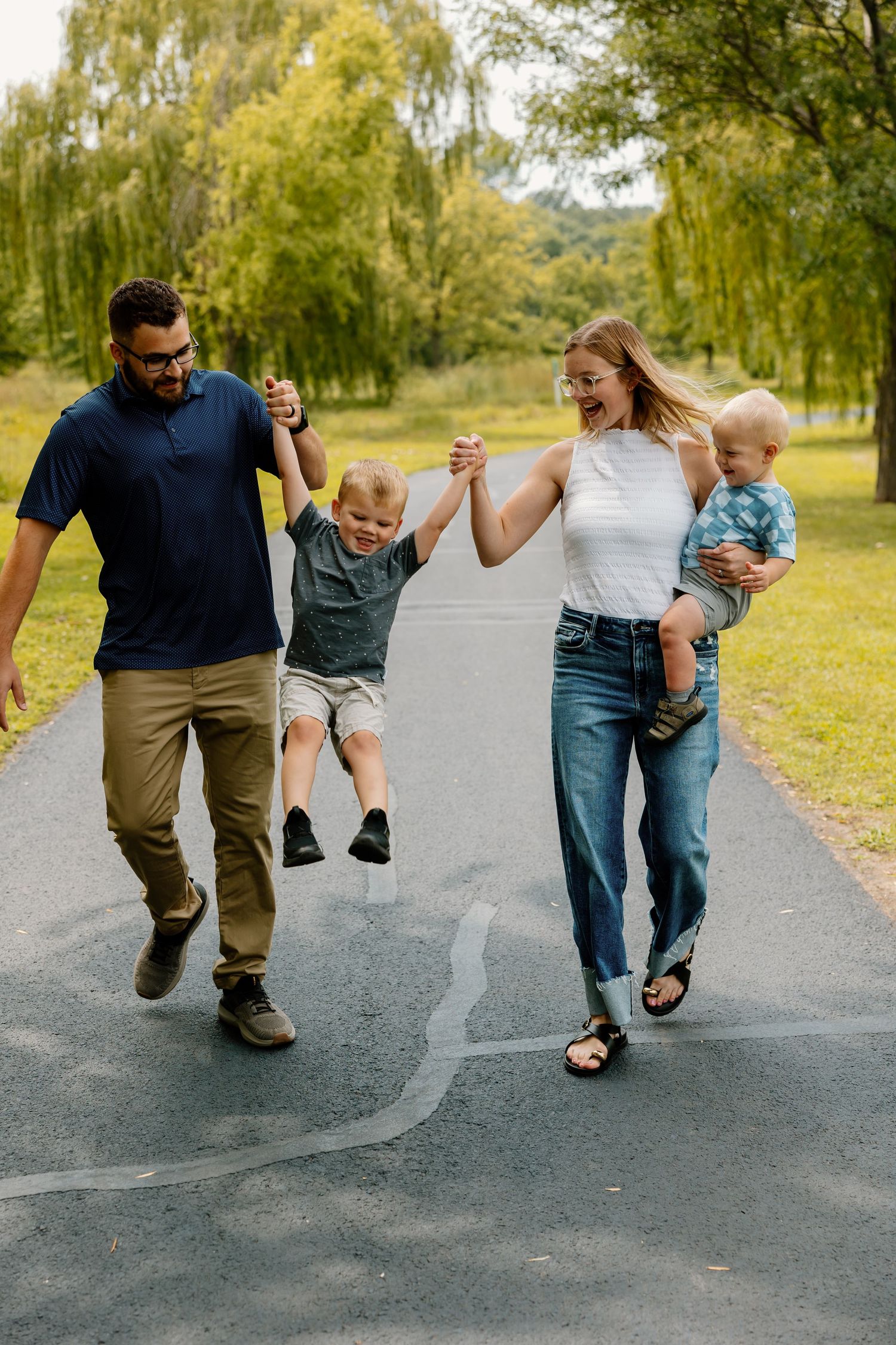Parents swing smiling toddler between them while walking on a paved path surrounded by willow trees in a park.