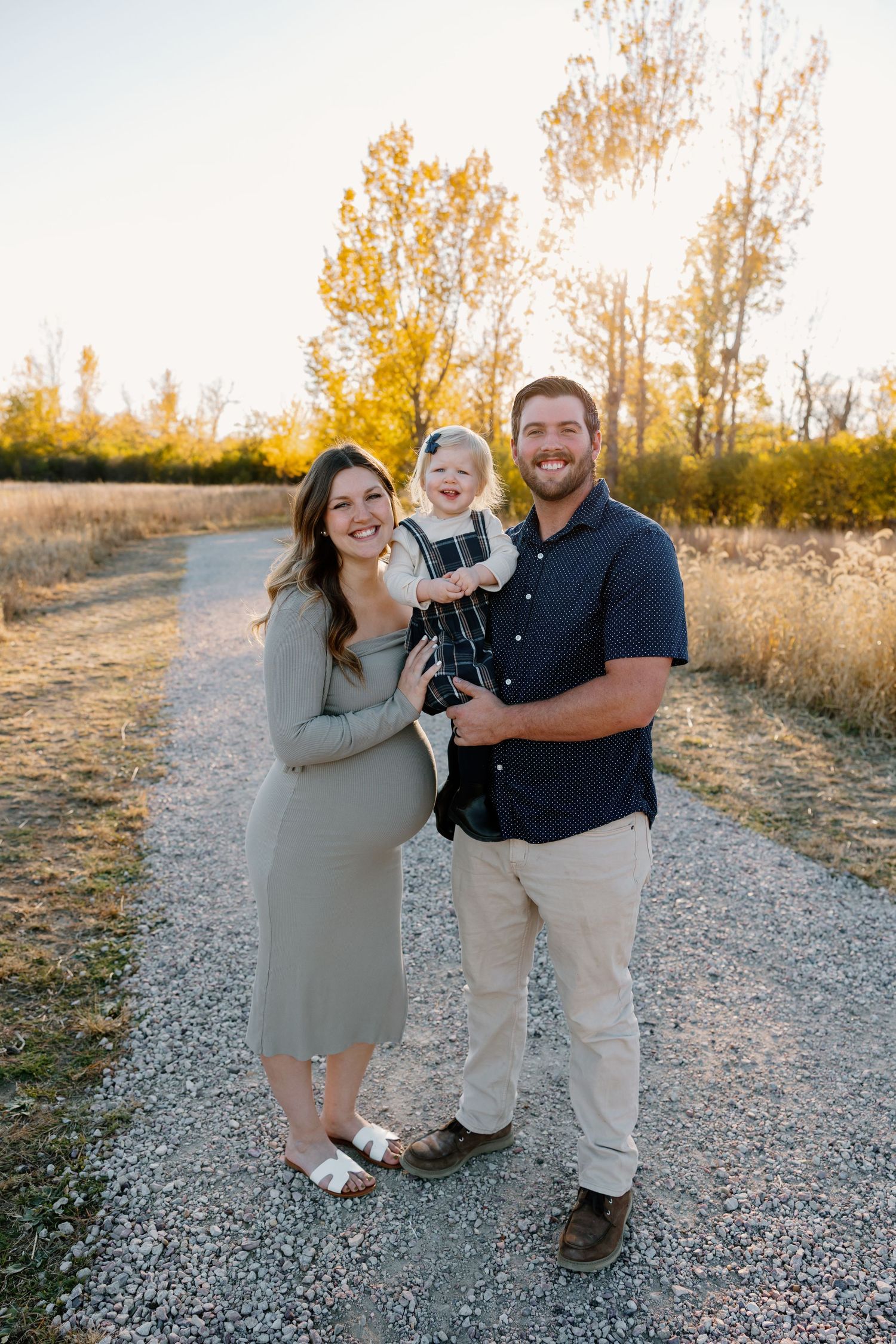 A family poses together on a gravel path during golden hour in autumn with trees in the background. Riley Ro Photo Sioux Falls, SD