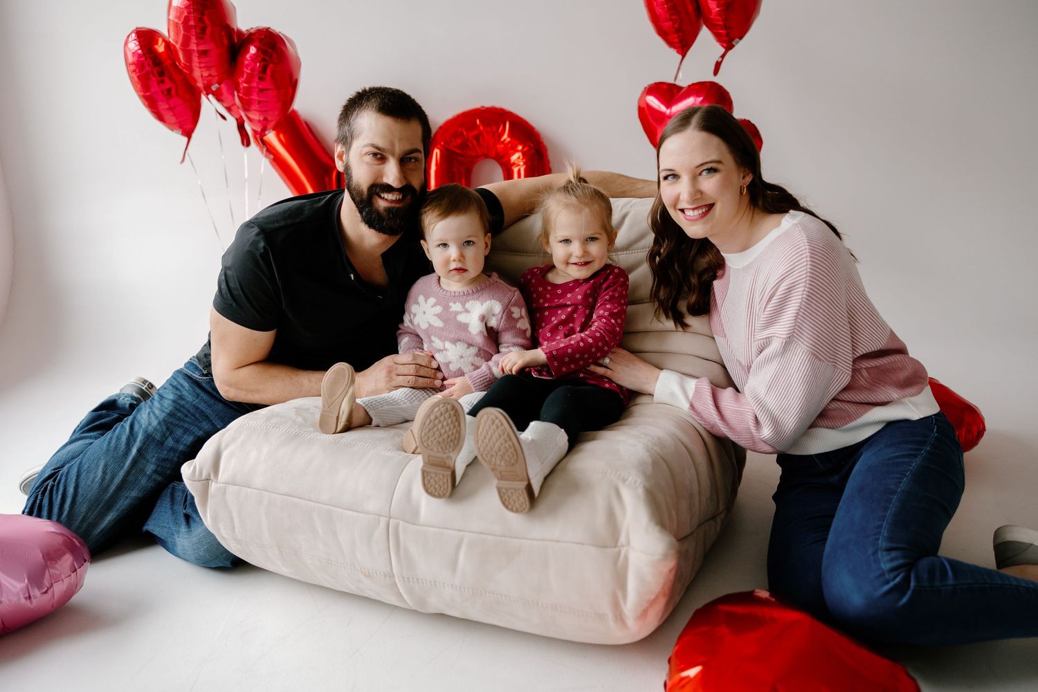 A family sits together on a beige couch with red heart balloons decorating the room for Valentine's Day.