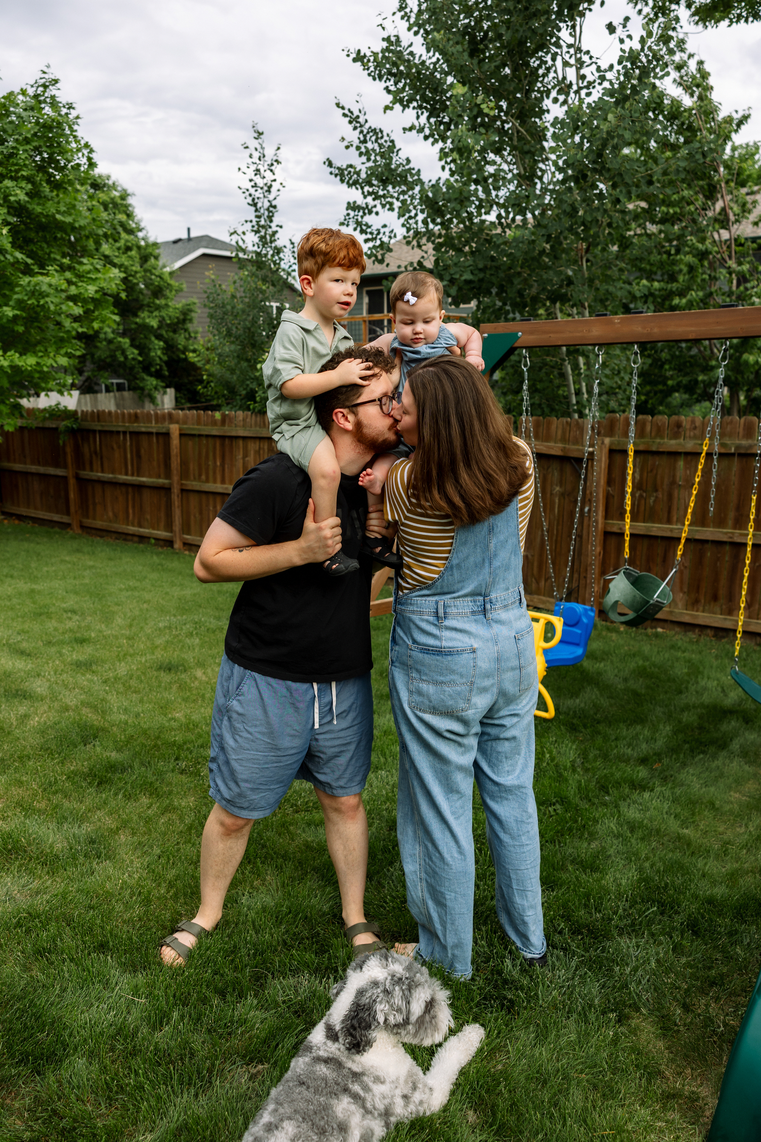 Family of four with dog playing in backyard near swing set on cloudy day.