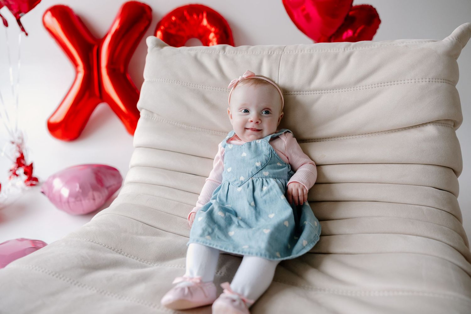 Sweet baby wearing light blue dress lying on beige couch with red balloon decorations in background.