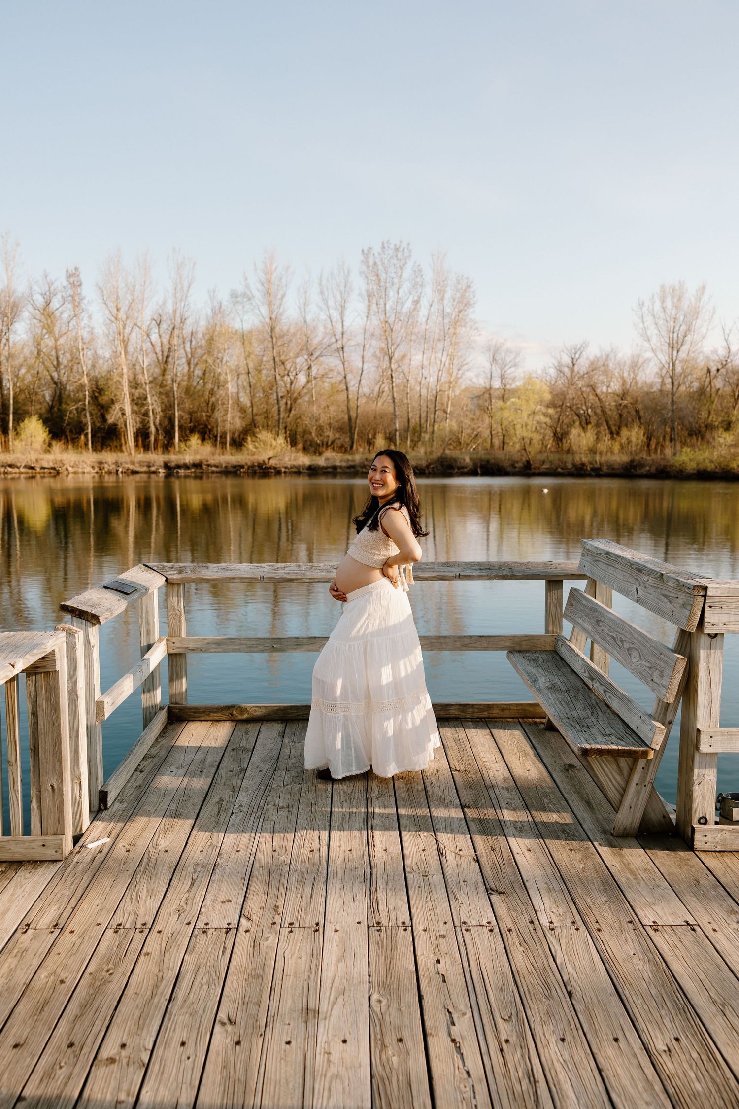 A figure in a flowing white dress stands on a wooden dock overlooking a serene lake surrounded by bare trees at sunset.