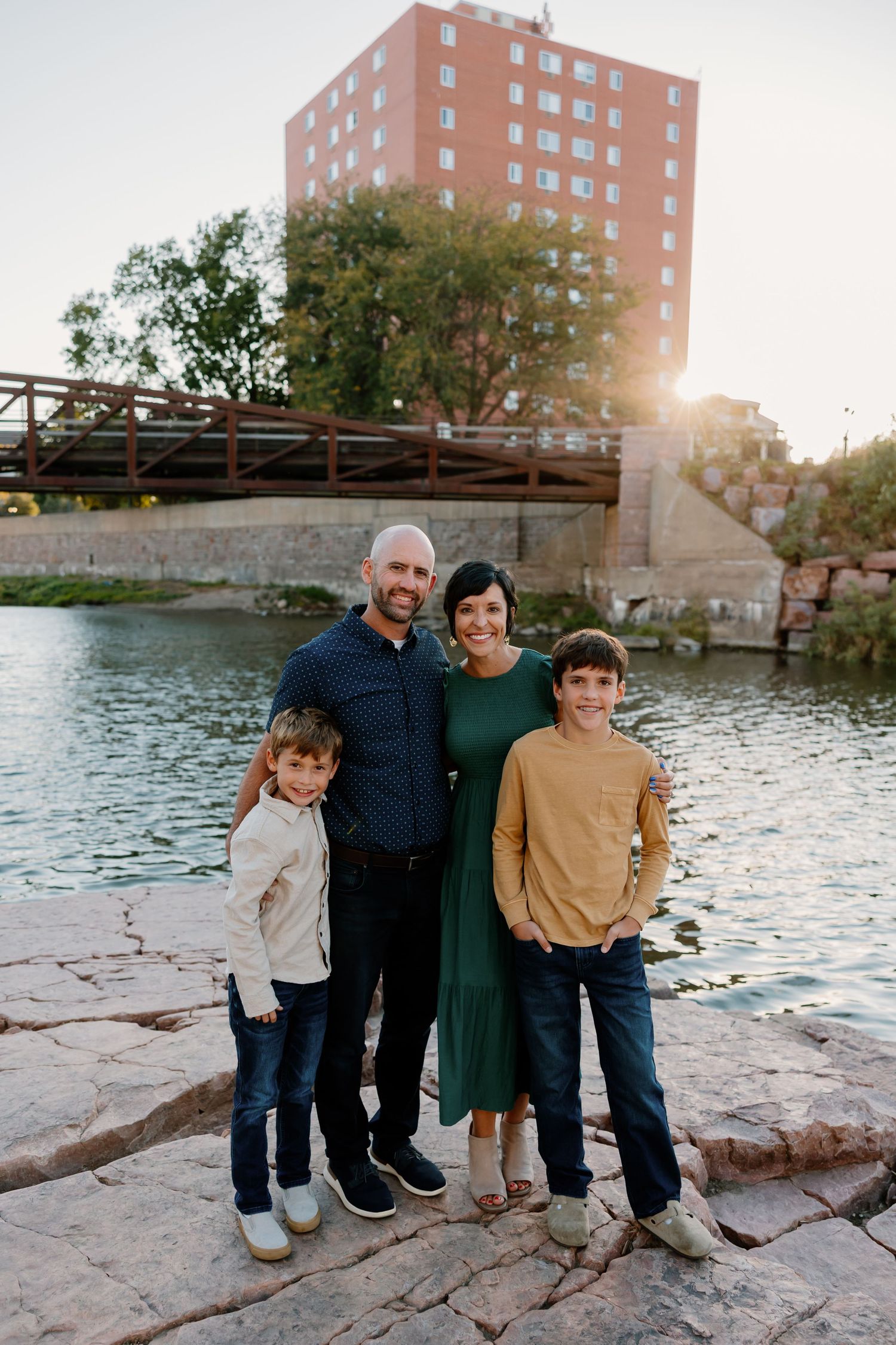 A family stands together on rocky terrain near a lake with a wooden bridge and brick apartment building in the background. Riley Ro Photo Si