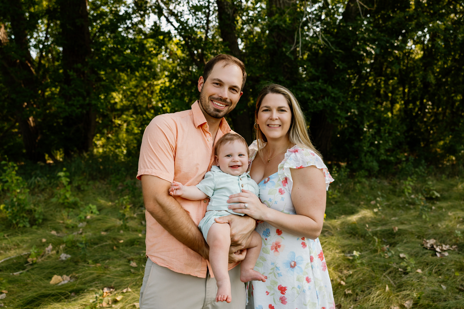 Happy young family with mother, father, and baby posing outdoors in a lush green park setting.