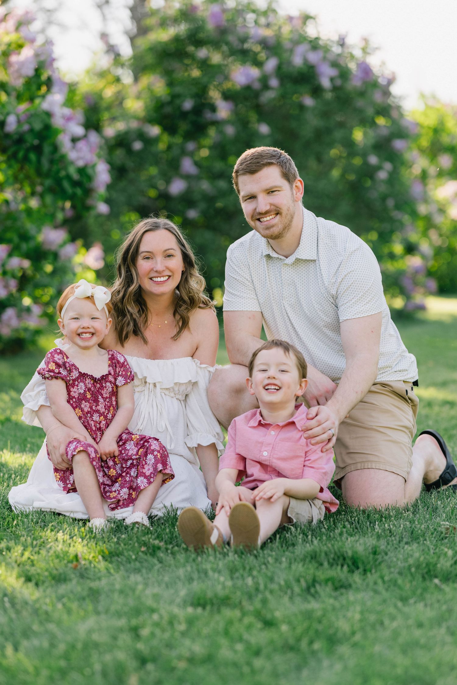 Happy family of four poses on green grass with blooming lilac bushes in a sunny outdoor portrait.