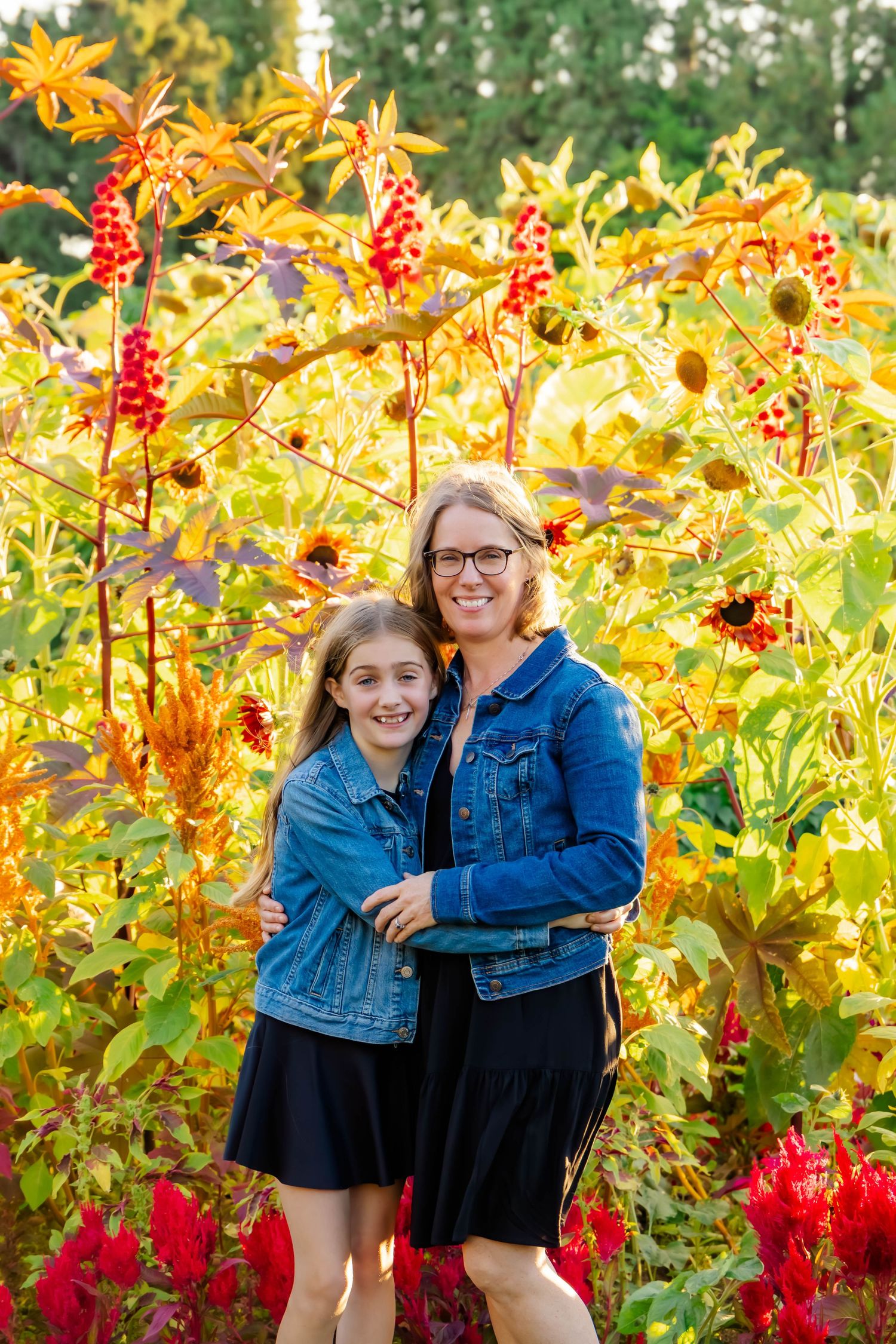 Two people in denim jackets pose together against a vibrant backdrop of yellow and red flowers in a garden setting.