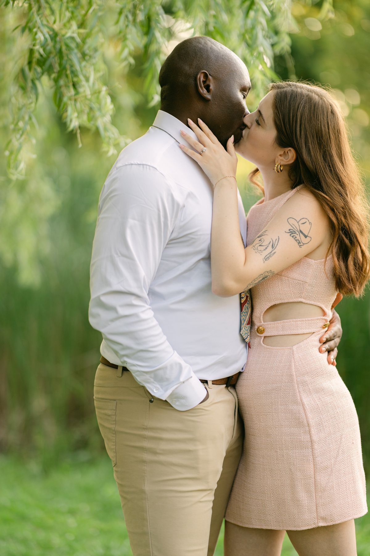 Interracial couple sharing a romantic kiss outdoors, surrounded by lush green foliage in a park setting.