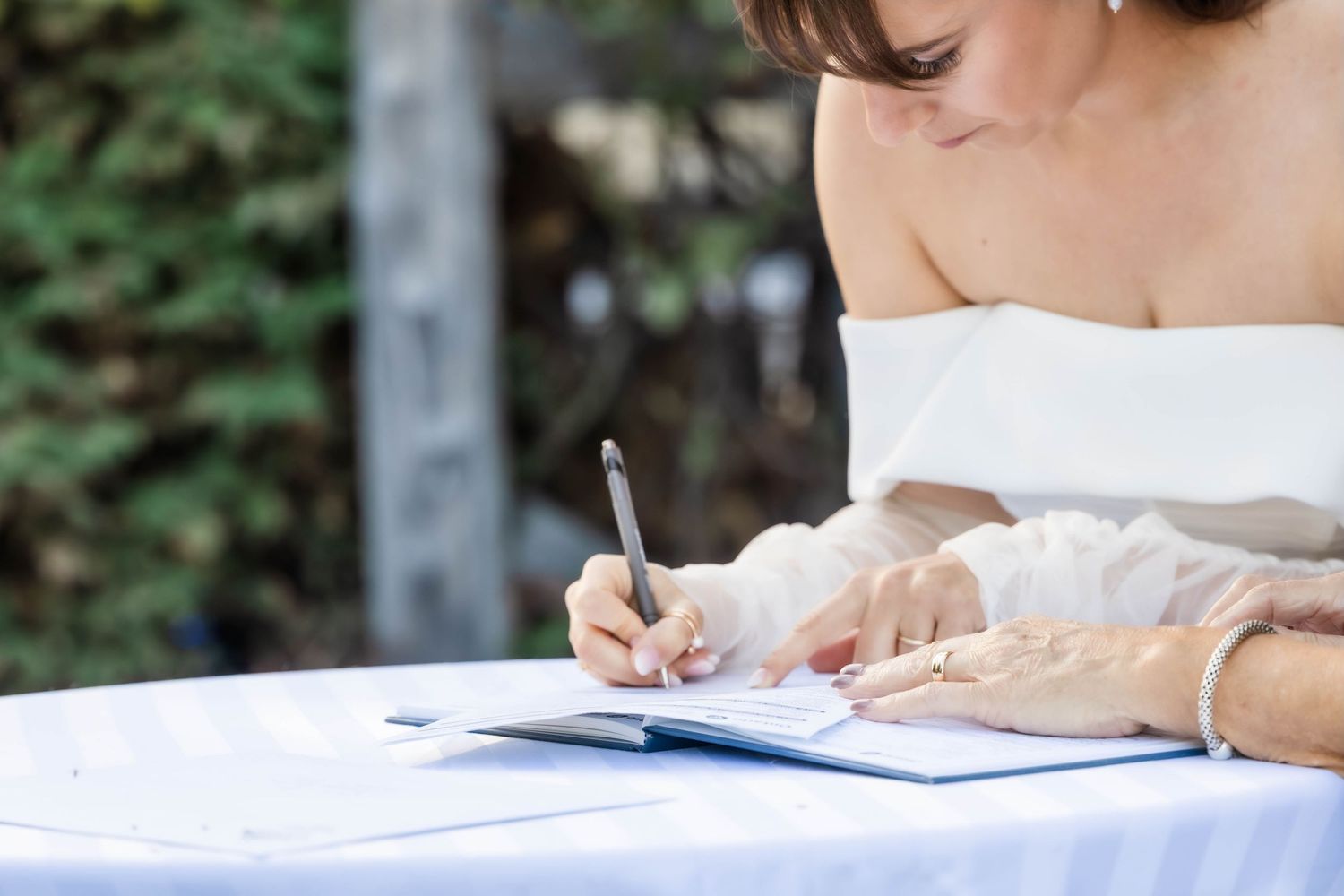 Bride in an off-shoulder wedding dress signs documents during an outdoor ceremony.