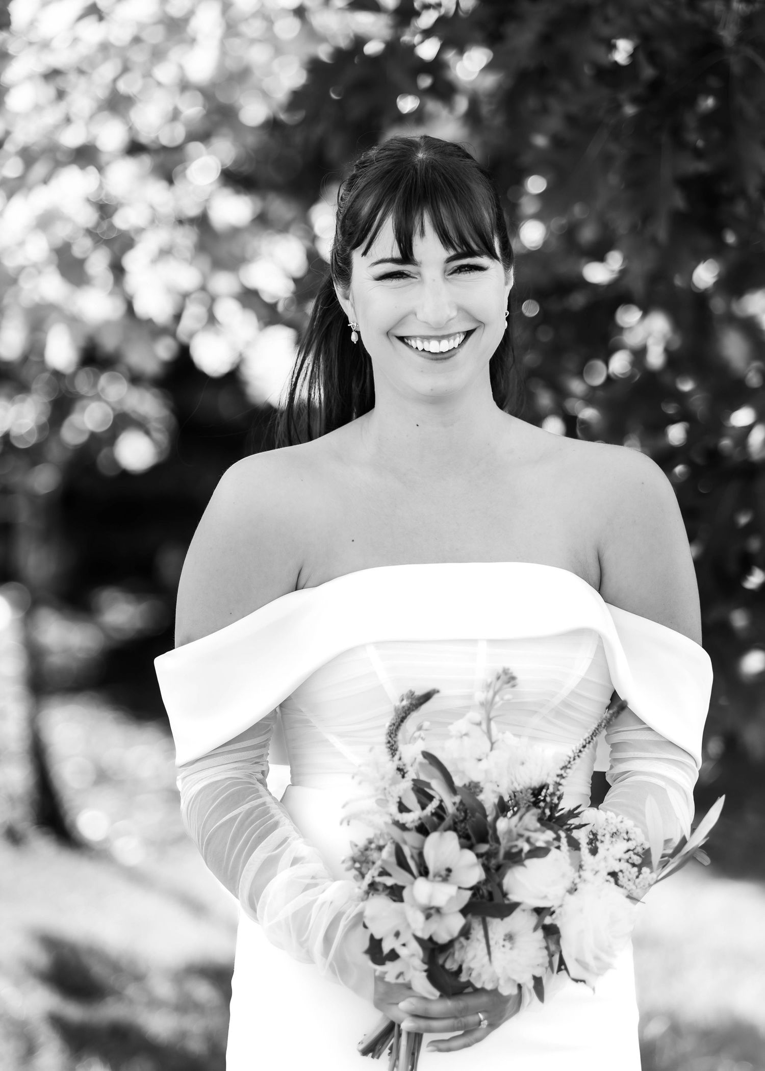 Elegant bride in off-shoulder wedding dress holding floral bouquet in black and white photography.