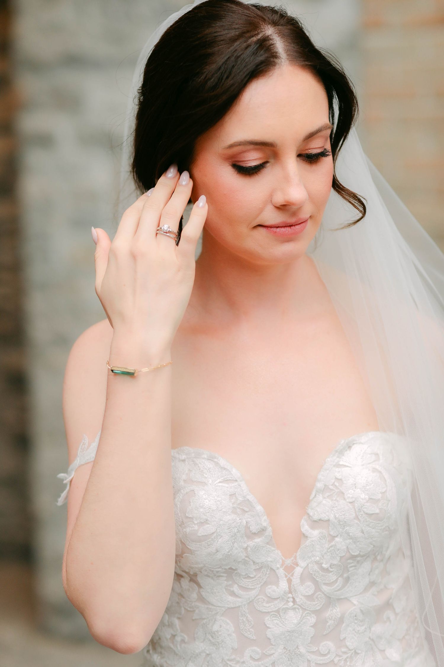 Elegant bride in lace strapless gown with veil, wearing a diamond ring, posing with hand near face.